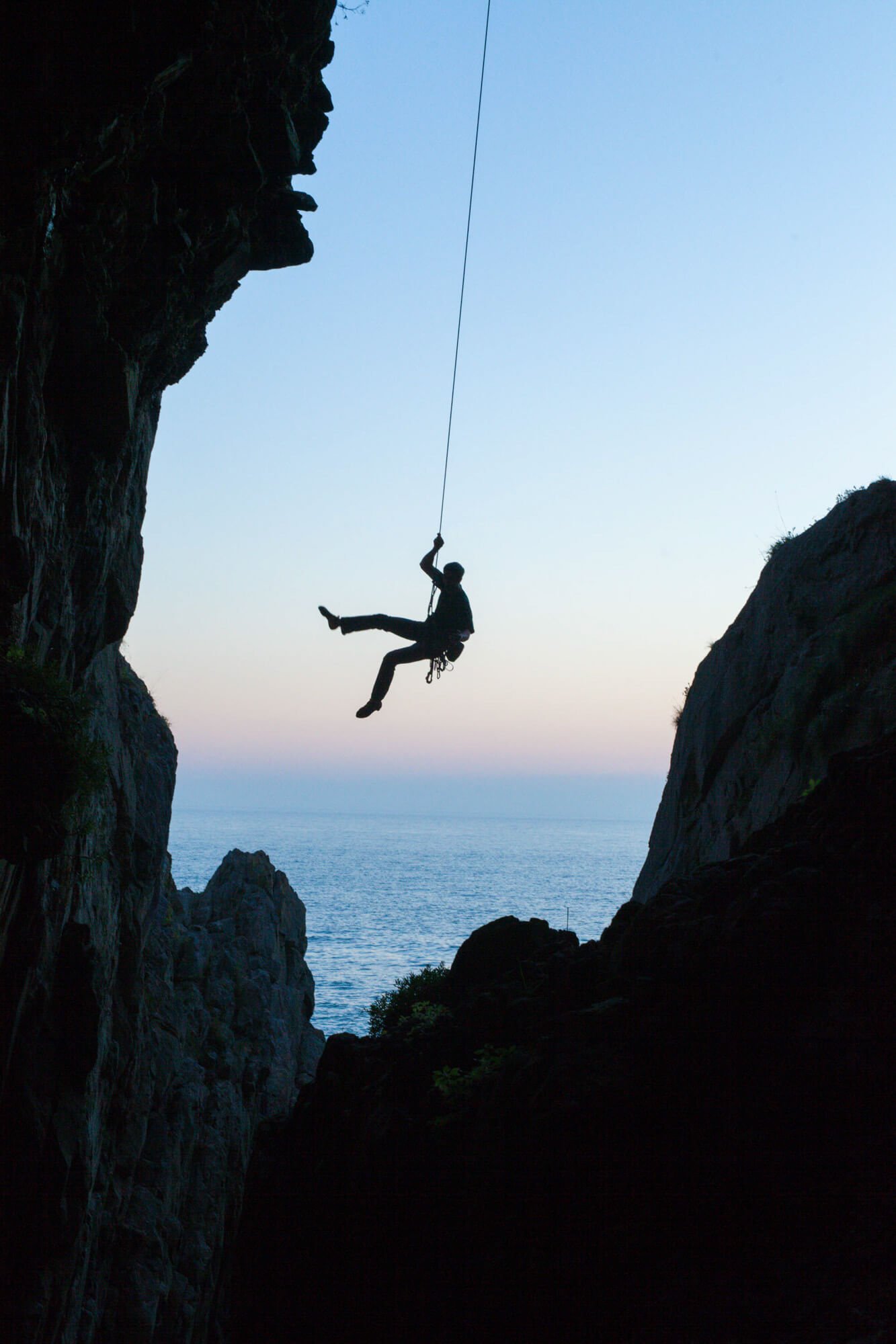 Silhouette of a person rappelling down a rock face against a sunset over the ocean.