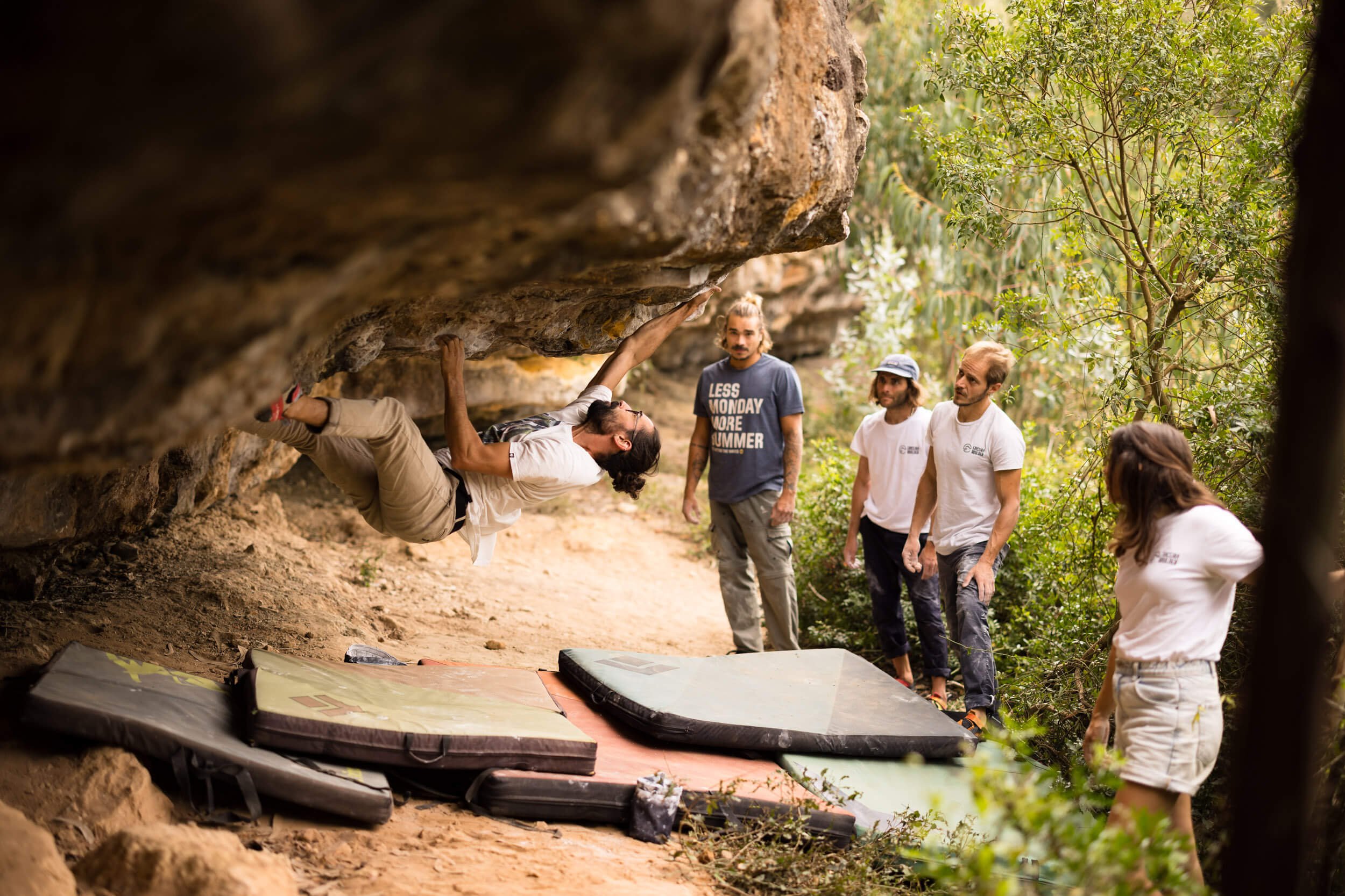 A person bouldering on a rock overhang with four spotters observing and multiple crash pads below for safety.