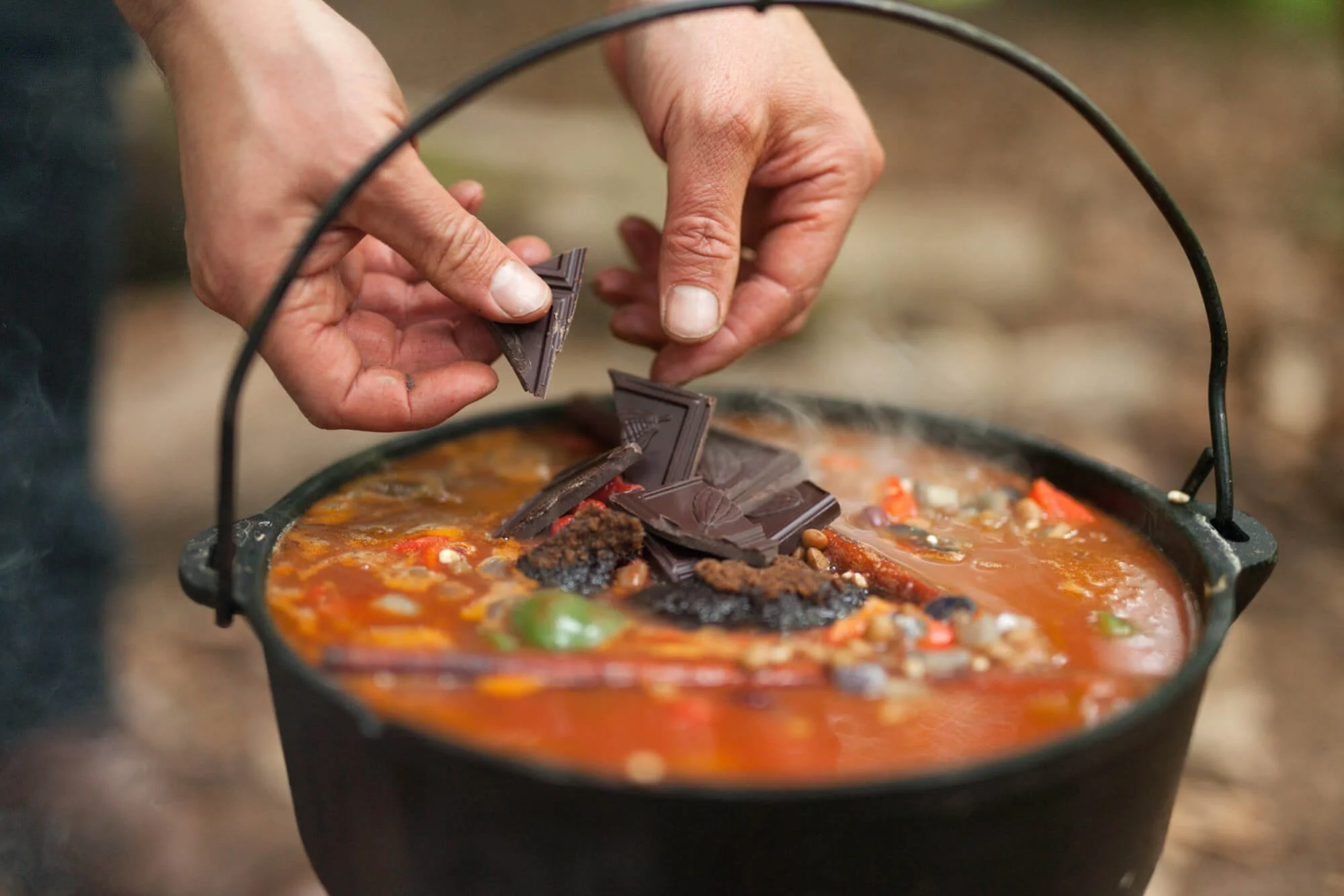 Person adding chocolate to a pot of chili or stew outdoors.