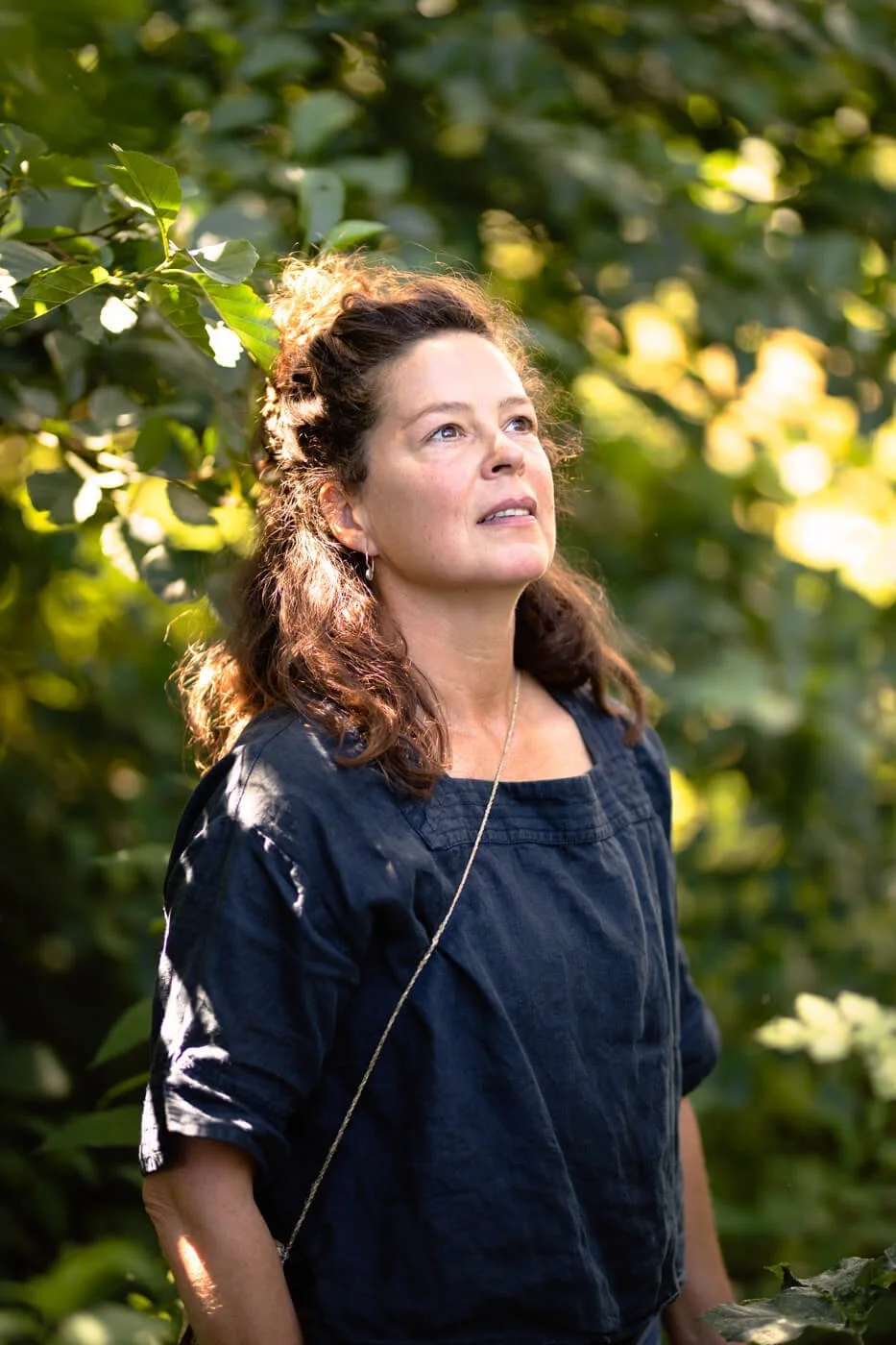A woman with long curly hair, wearing a dark top, stands in a sunlit garden or forest area with lush greenery. She is looking upwards with a thoughtful expression.