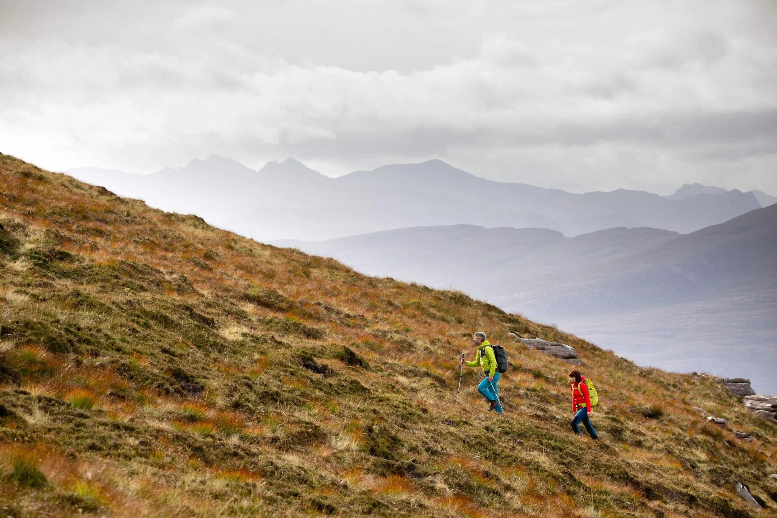 Two hikers ascending a grassy hillside with mountains in the background under a cloudy sky.