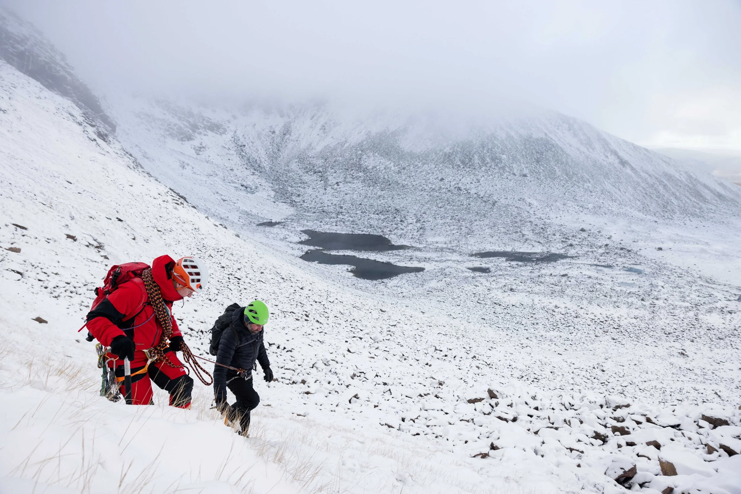 Two climbers in winter gear ascending a snowy mountain, with a foggy landscape in the background.