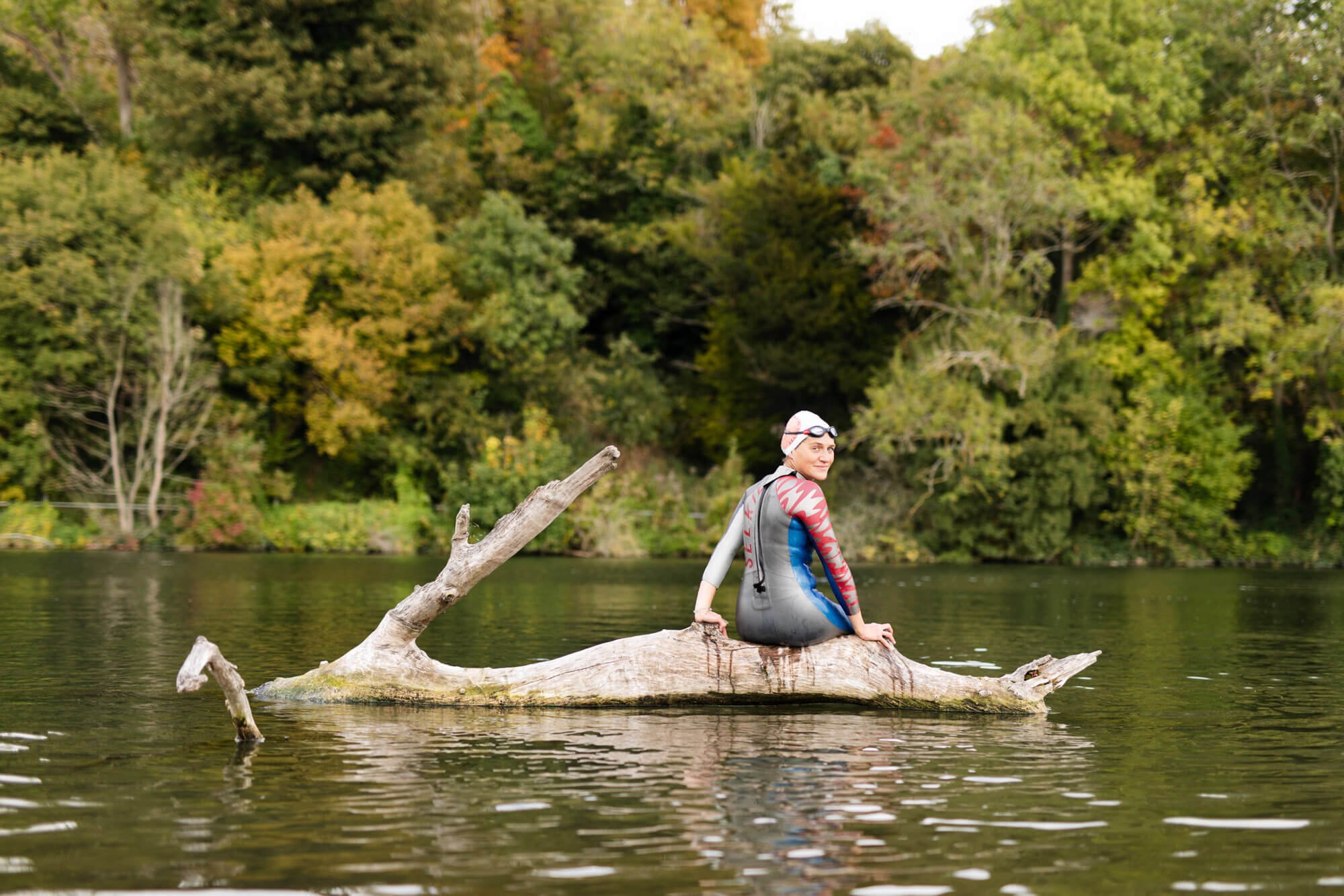 Person in a wetsuit sitting on a log in a lake surrounded by trees.