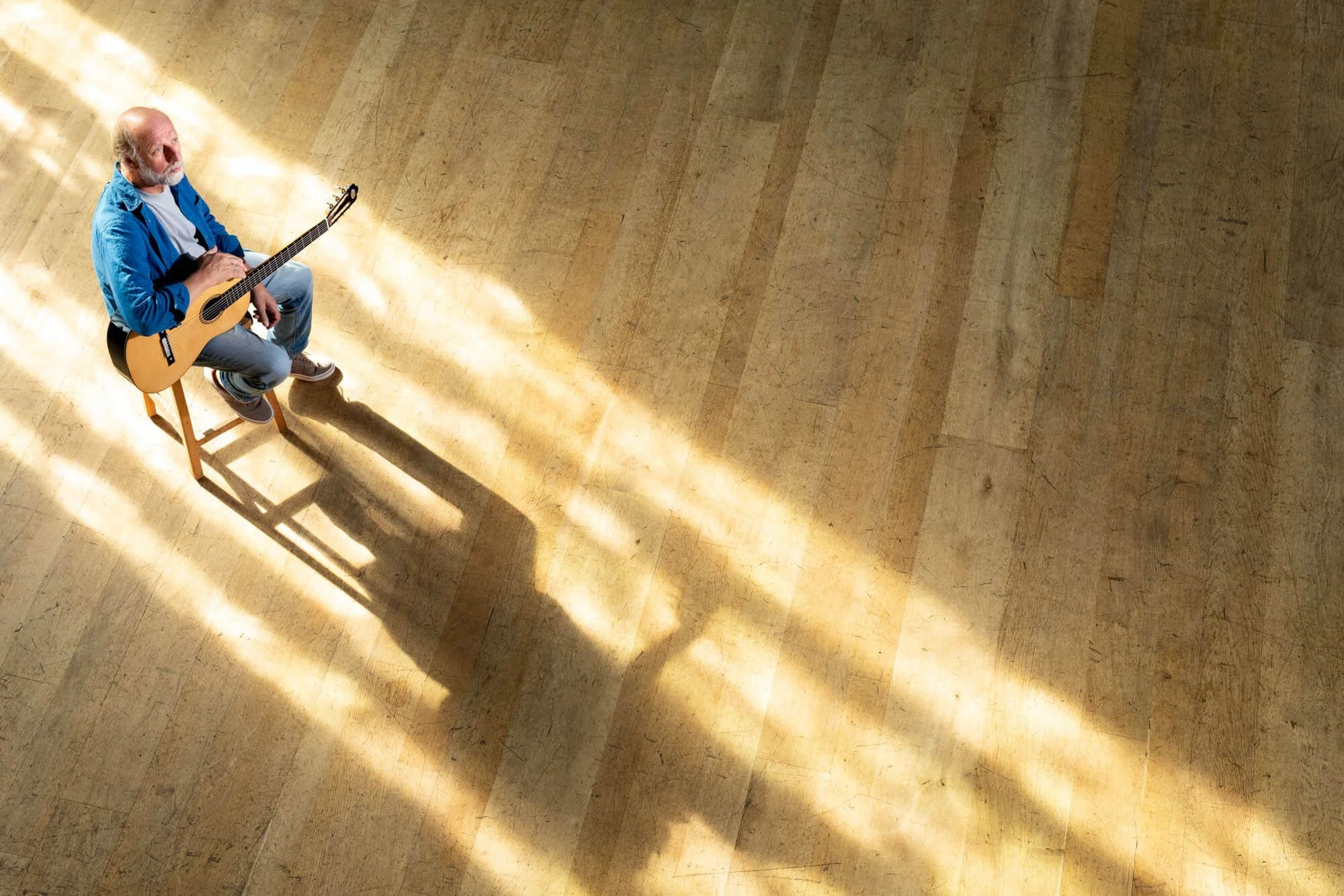 Man sitting on a chair playing guitar with sunlight casting shadows on a wooden floor.