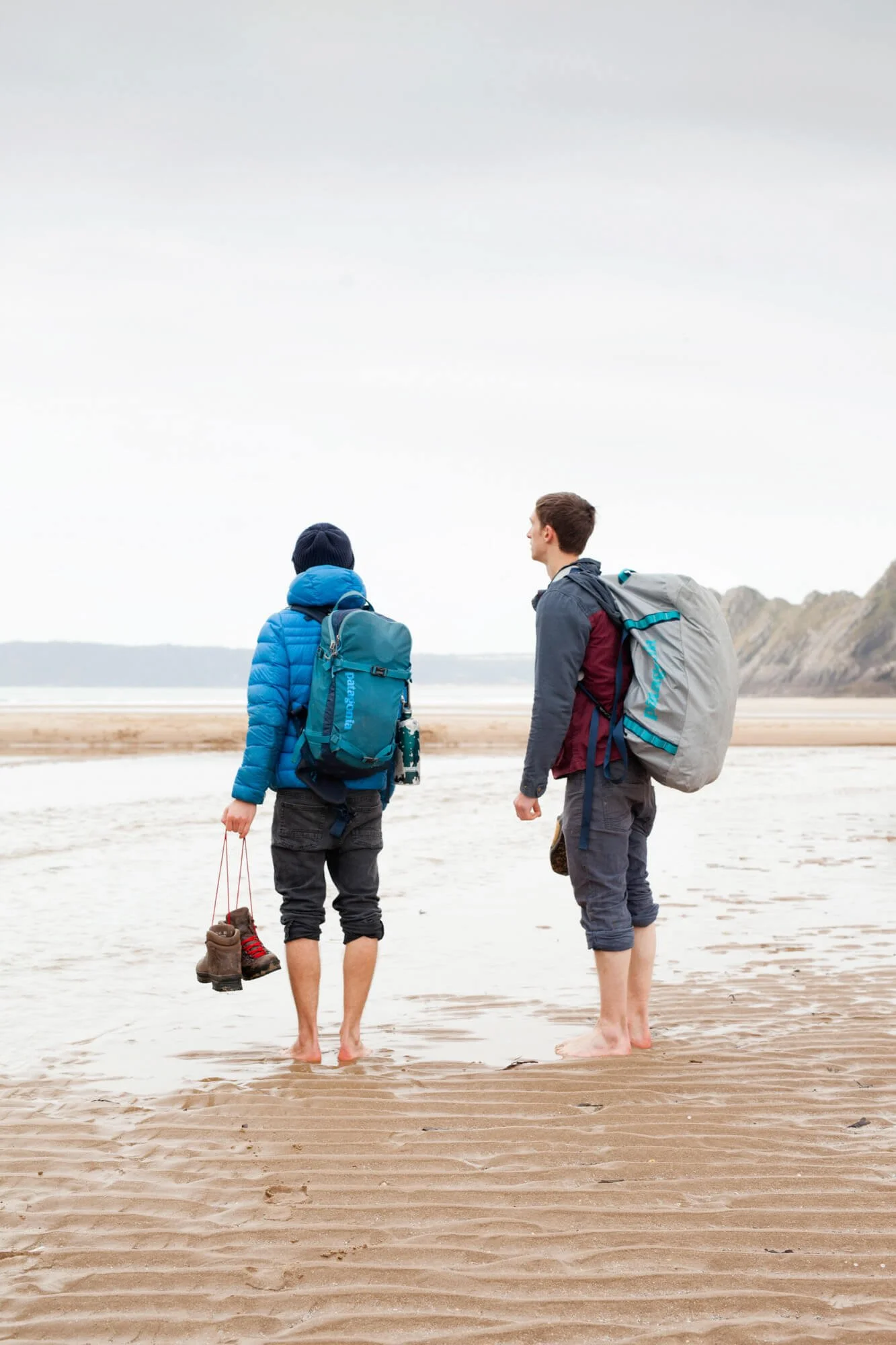 Two people with backpacks walking barefoot on a sandy beach holding shoes, facing the ocean.