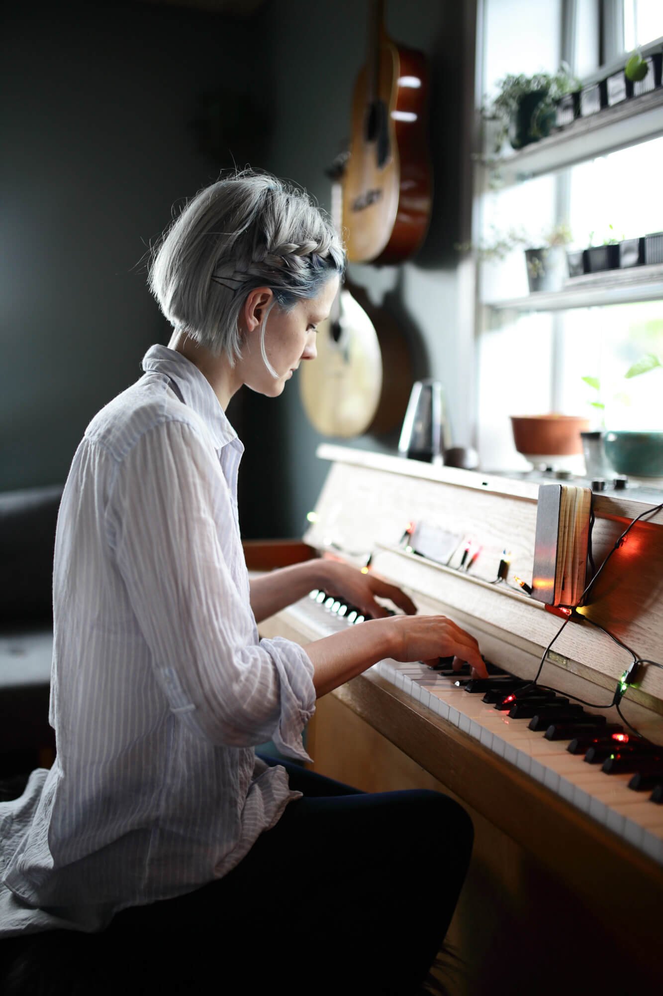 A person with short hair playing an upright piano in a room with guitars on the wall and plants on the windowsill.