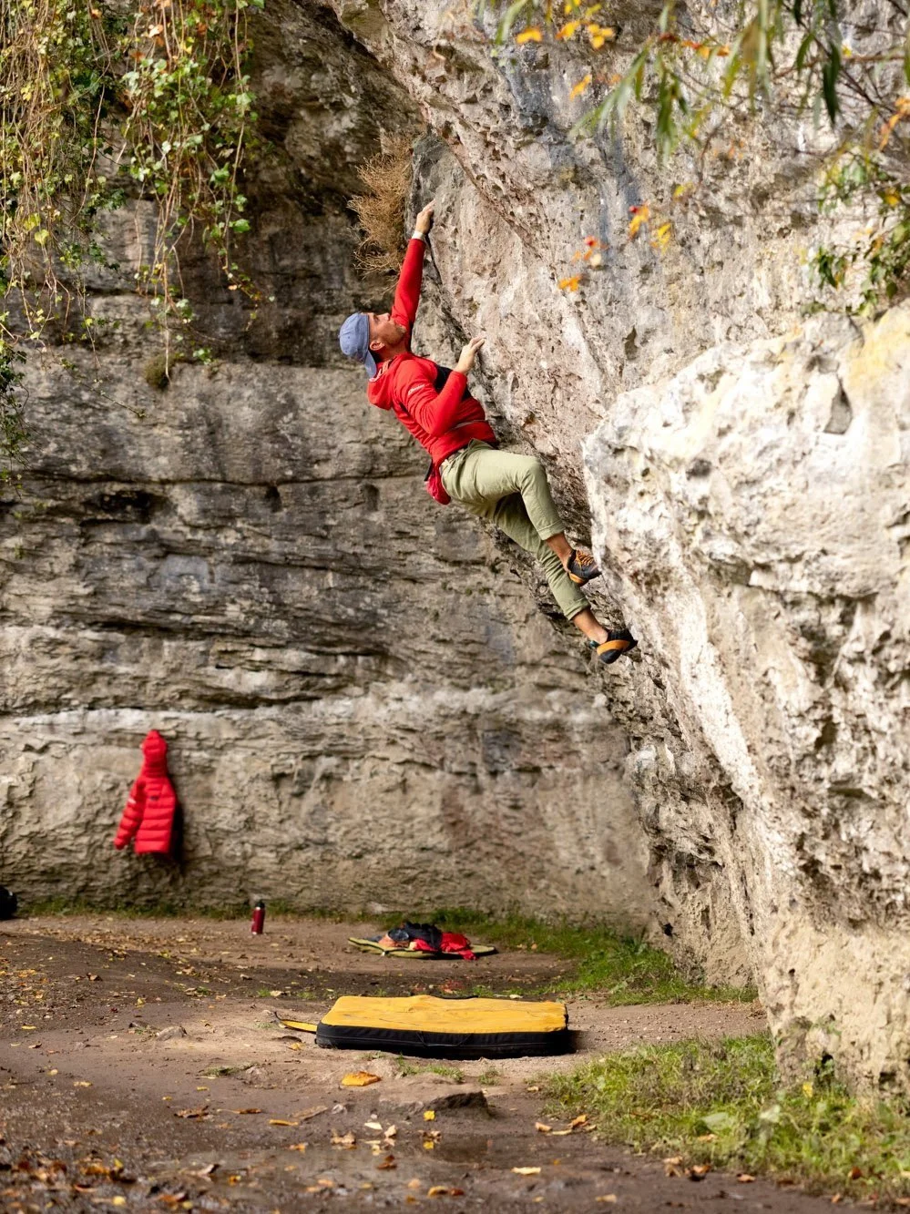 Climber Leo Houlding in red sweater and green pants scaling a rock face outdoors, with a blue cap and safety pad on the ground below.