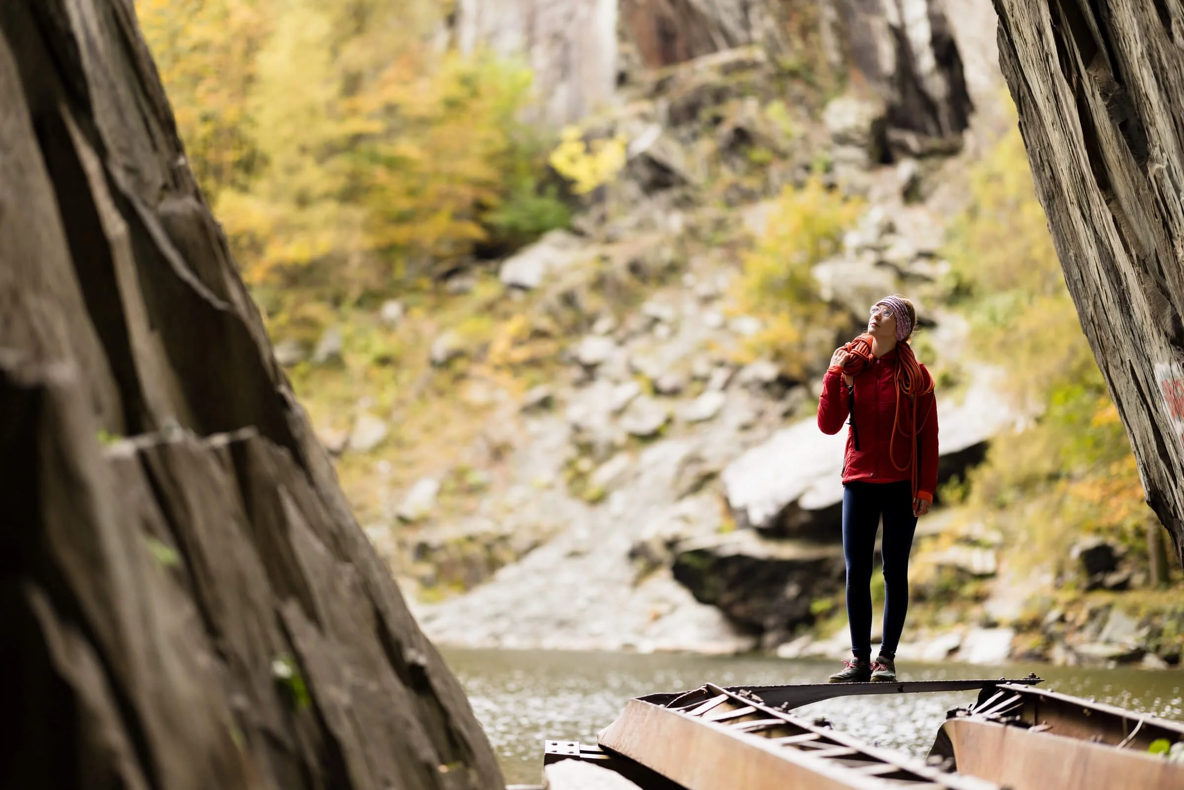 Person in red jacket holding climbing rope standing on a rocky platform near a cliff and water.