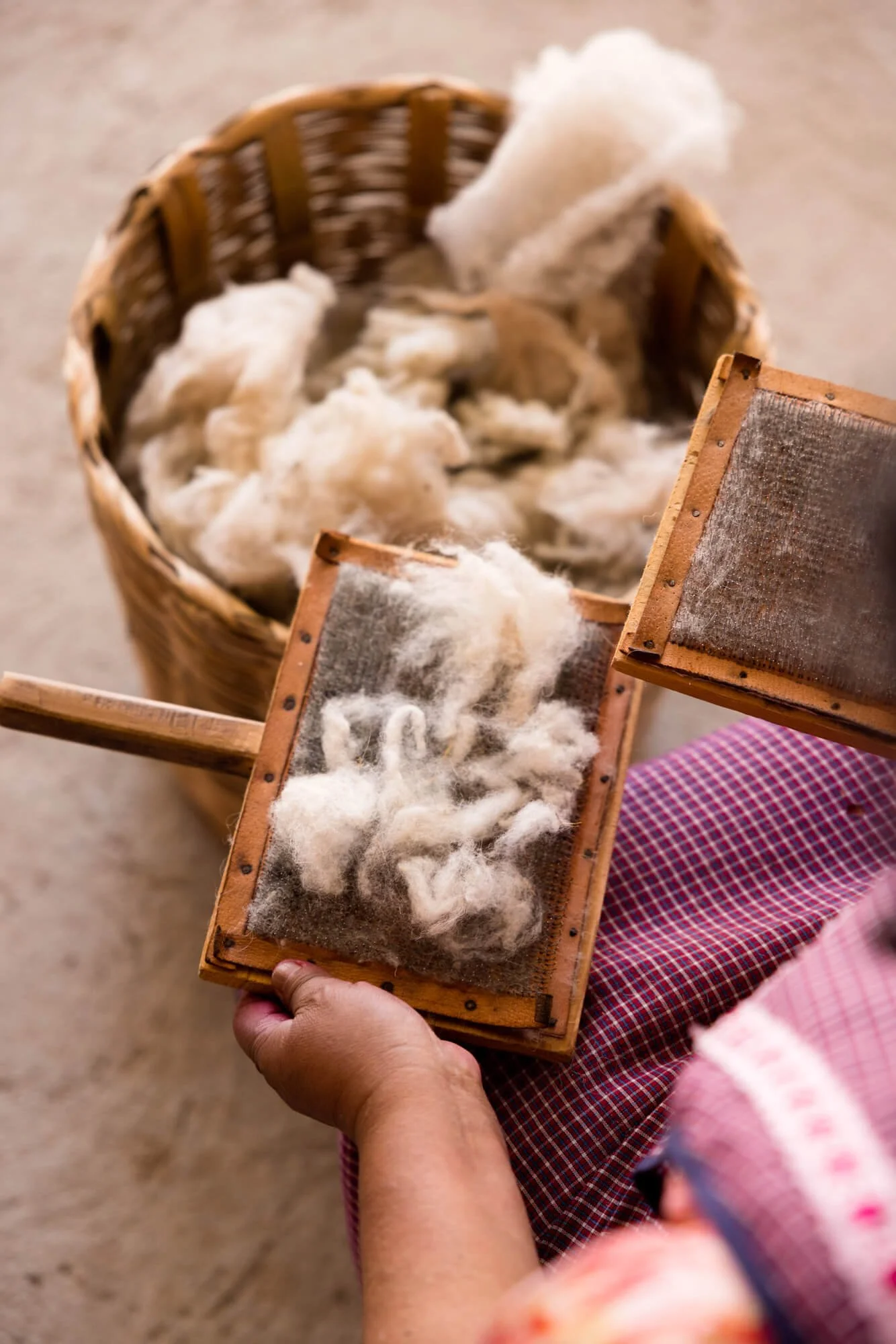 Woman using carding tools to process wool near a basket full of wool fibers.