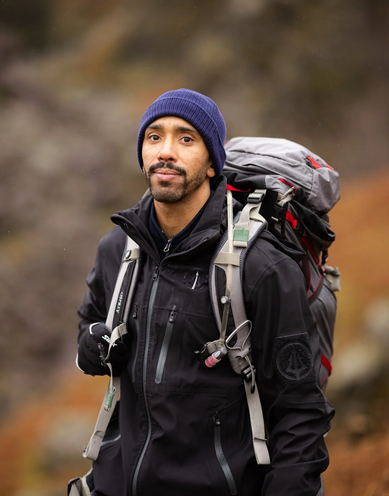 Man wearing a blue beanie and black outdoor jacket with a backpack, standing outdoors.