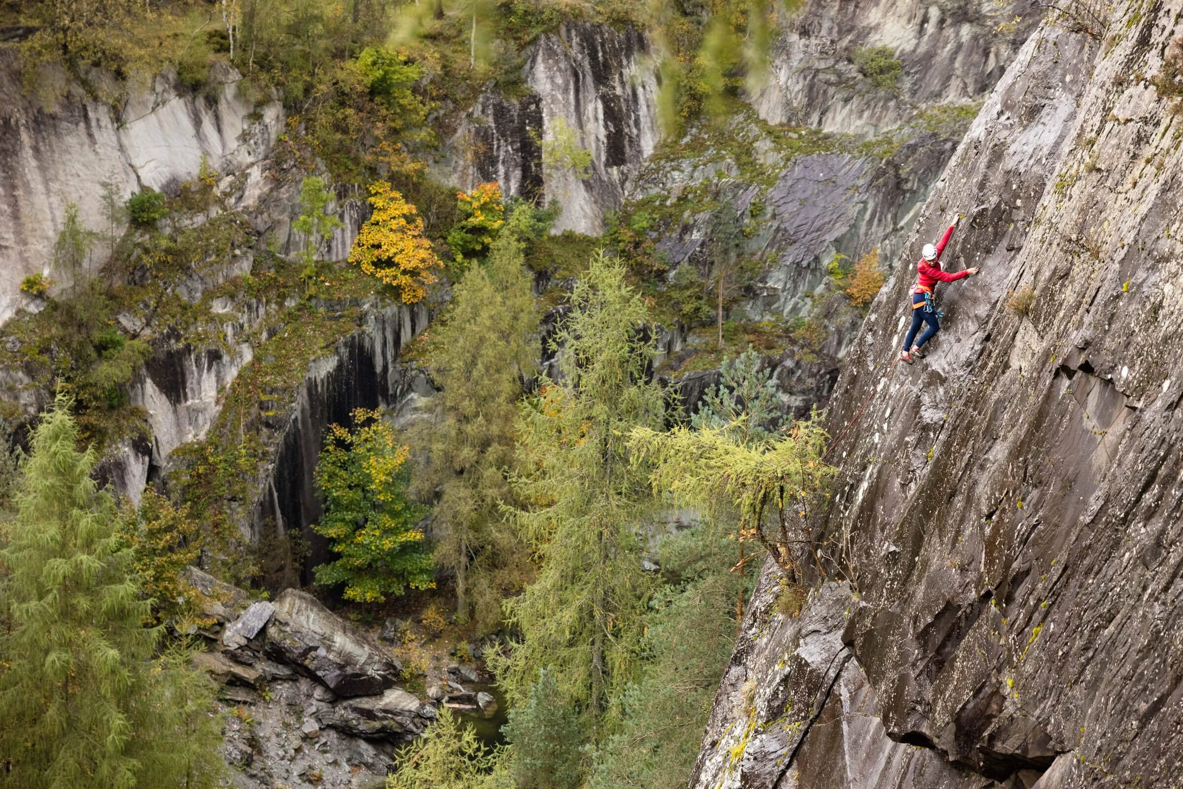 Rock climber ascending a steep rock face in a forested area with autumn foliage.