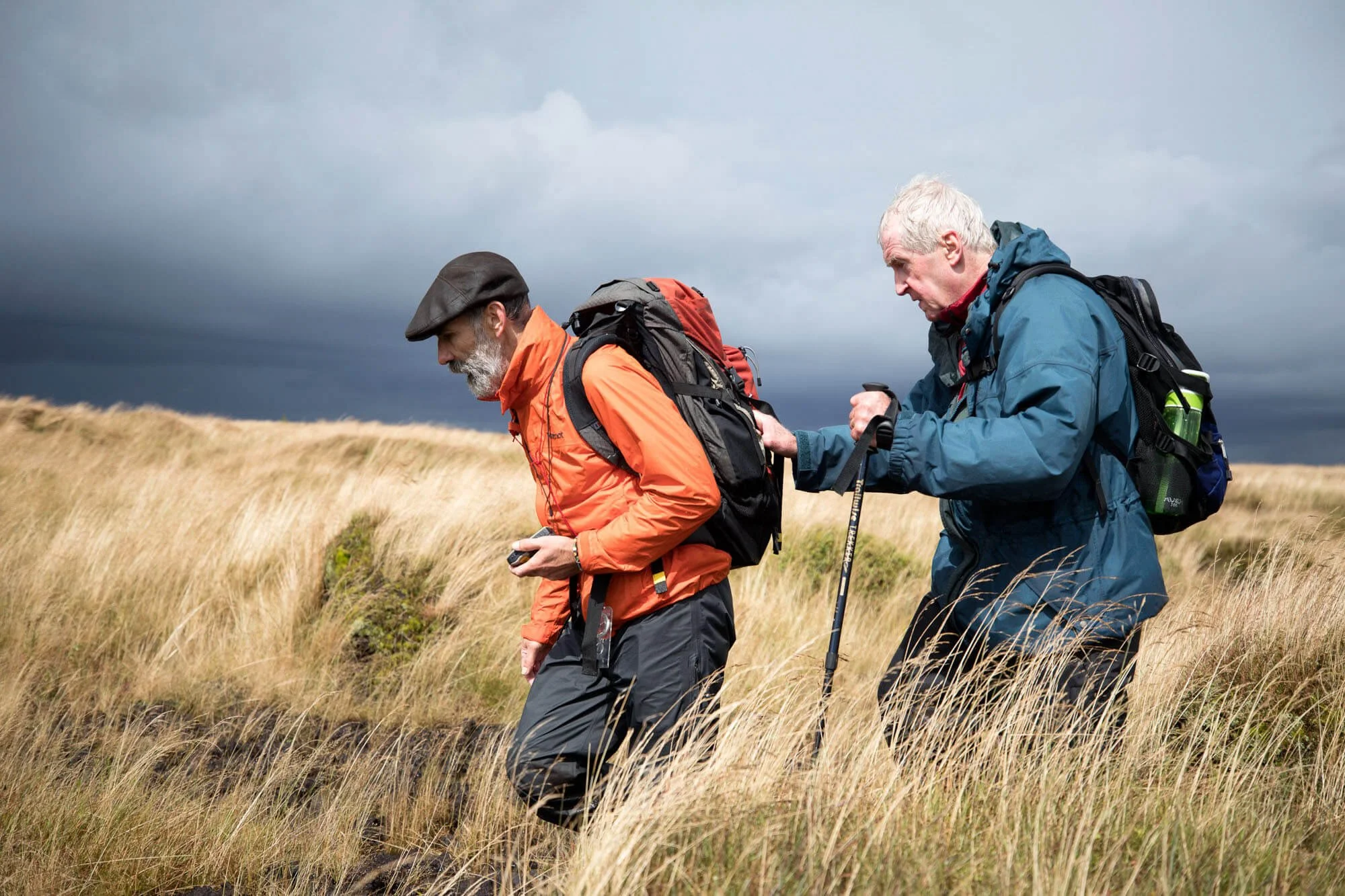 Two hikers walking in a grassy field, one using a walking stick, both wearing outdoor gear and backpacks, with stormy skies in the background.