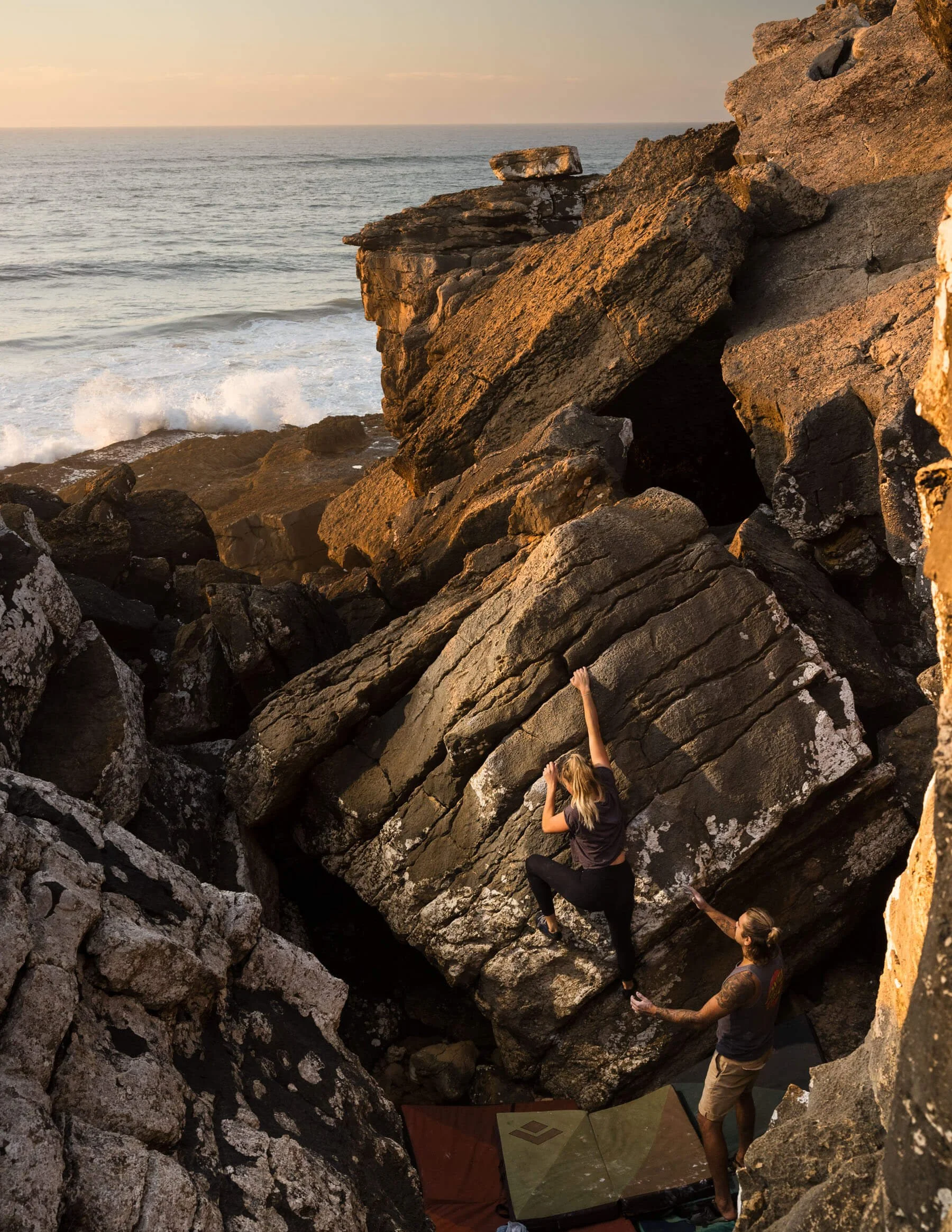 A person rock climbing on a large boulder near the ocean with a spotter below and crash pads on the ground. The sea is visible in the background with waves crashing against the rocks during sunset.