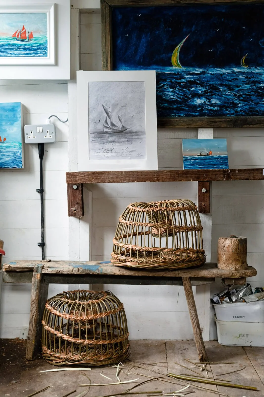 Art workshop with paintings of sailboats, wicker traps, and a rustic bench.