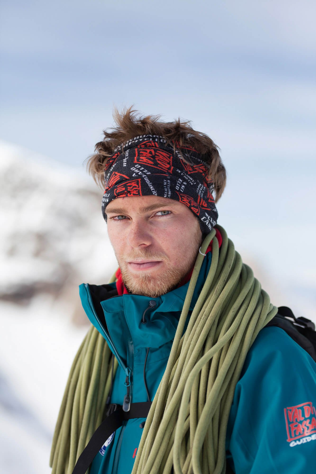 Man wearing a headband and teal jacket with climbing rope on shoulder, standing in snowy landscape.
