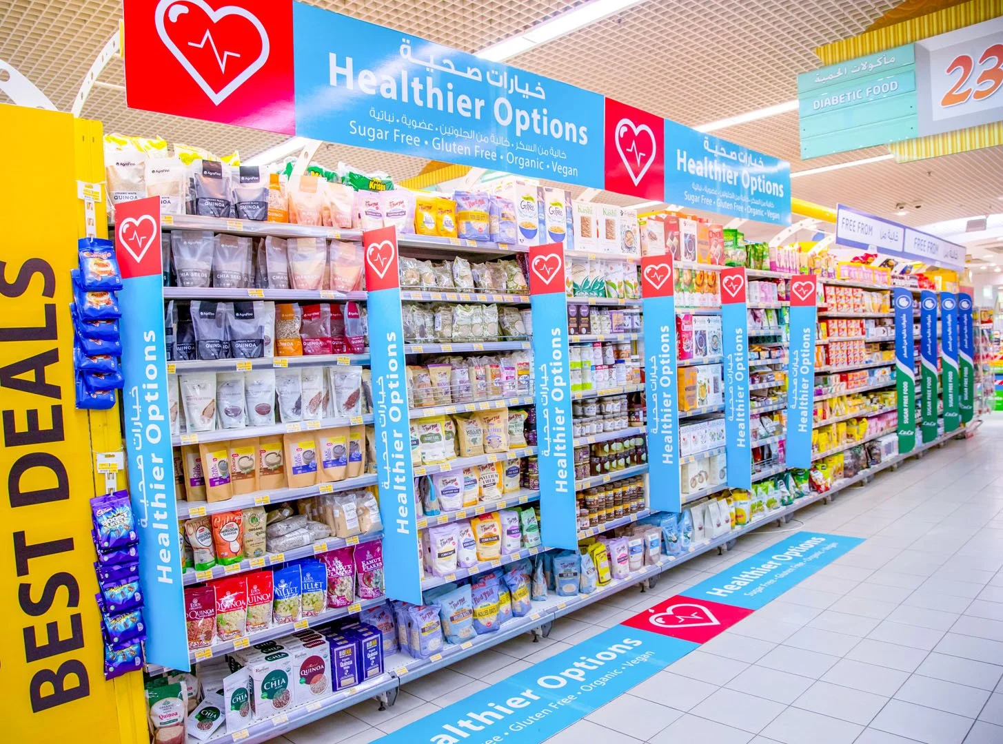 A grocery store aisle with shelves stocked with health-focused products like sugar-free, gluten-free, organic, and vegan options, under a blue sign that reads 'Healthier Options'.