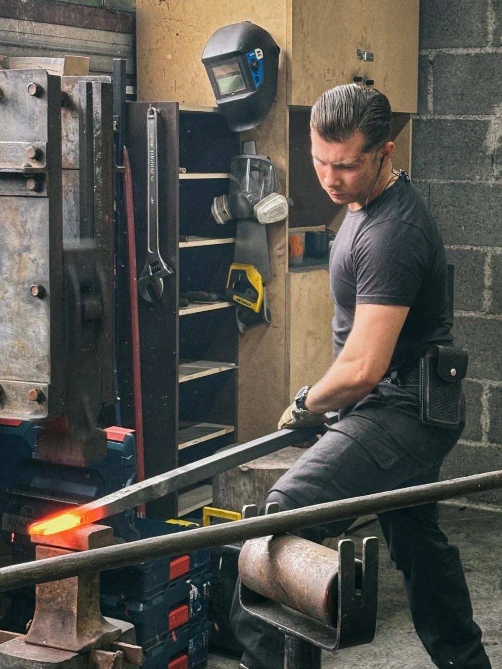 Un homme travaillant dans un atelier de forgeron, utilisant une barre de métal rougeoyant, avec divers outils de protection et un masque de soudure suspendu en arrière-plan.