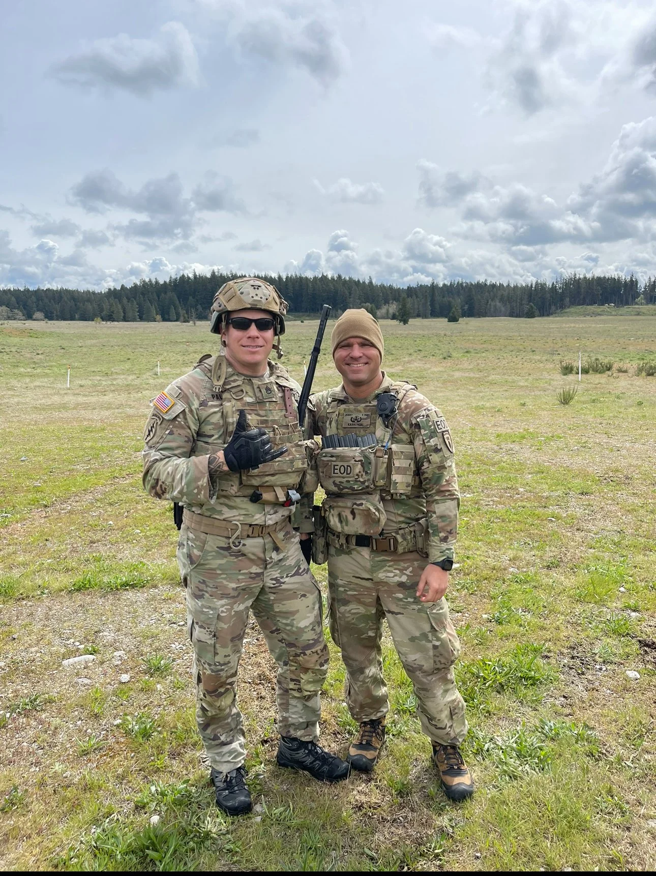 Two soldiers in camouflage uniforms standing on an open field, smiling at the camera, with one making a finger gun gesture. Overcast sky with clouds and trees in the background.