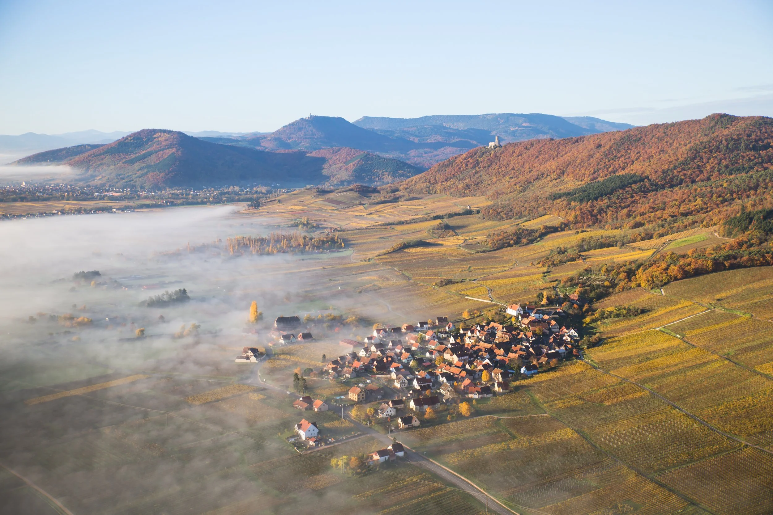Vue aérienne d'un village entouré de terrains agricoles et de montagnes avec de la brume matinale dans la vallée.