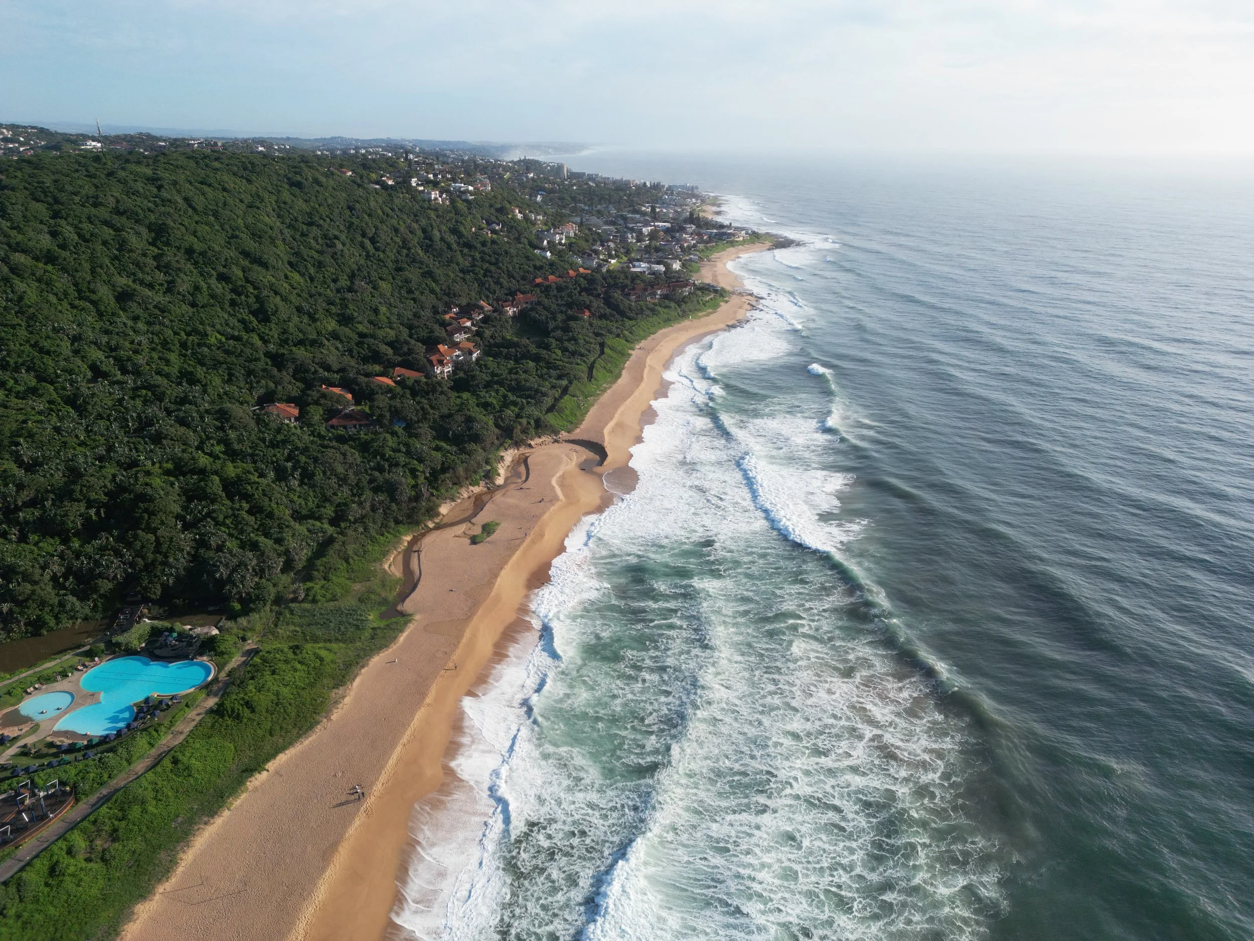Aerial view of a coastline with sandy beach, ocean waves, lush green hillside with houses, and a swimming pool nearby.