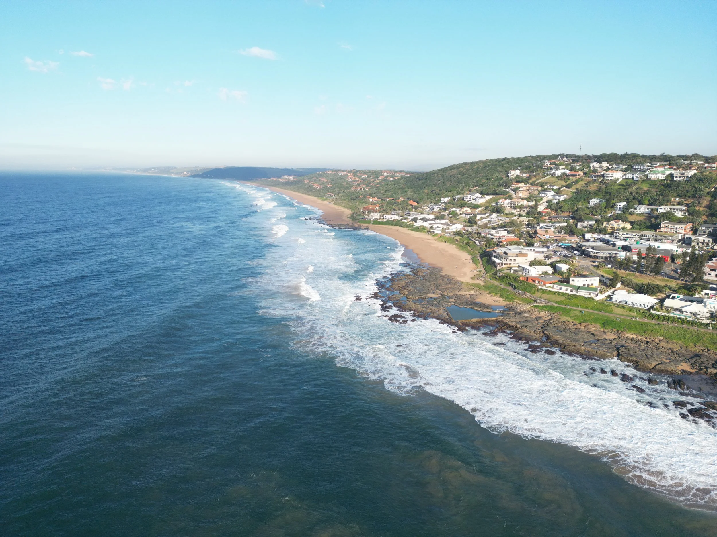 An aerial view of a coastal city with a sandy beach, rocky shoreline, and residential buildings on a green hillside under a blue sky.