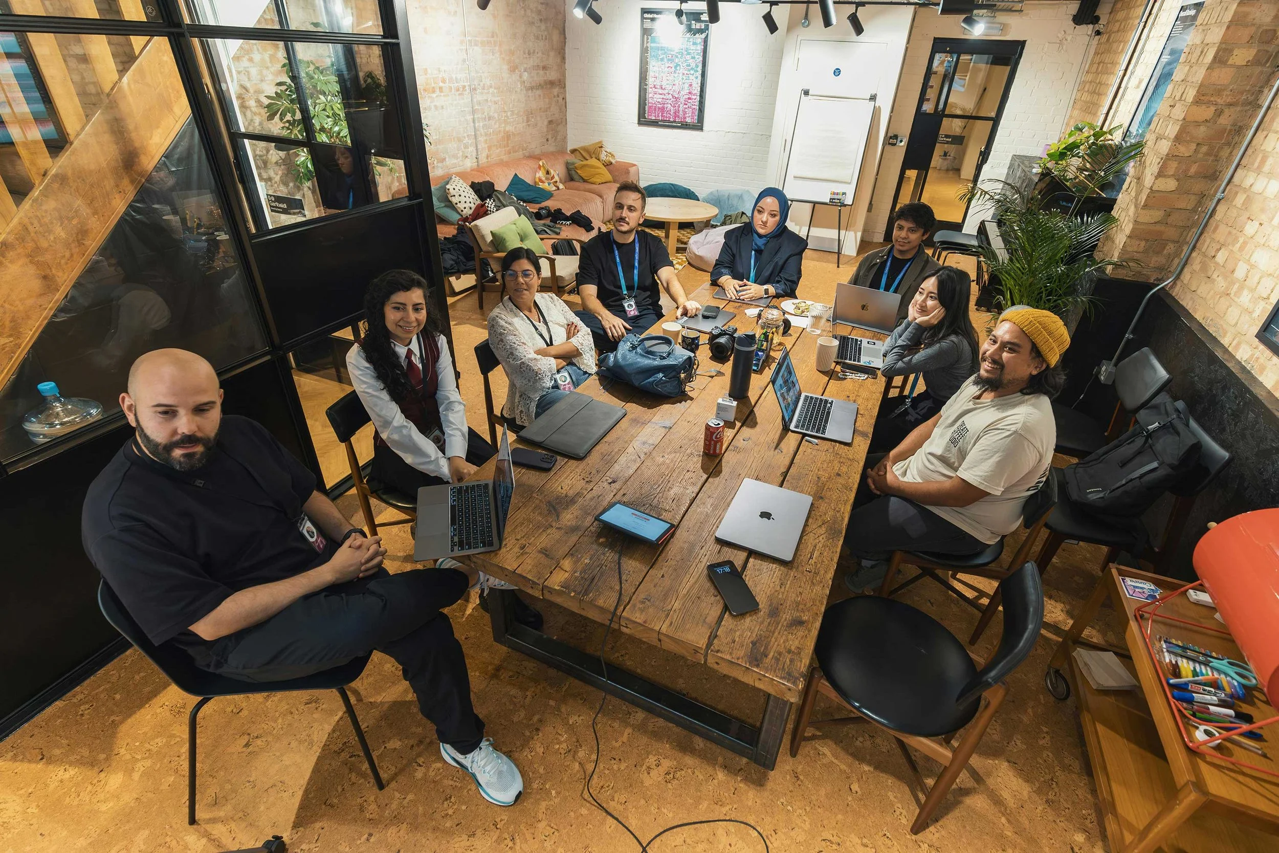 Groupe de personnes assises autour d'une grande table en réunion dans un espace de coworking ou de bureau à l'intérieur, avec des ordinateurs portables, des appareils photo, et d'autres objets sur la table, dans un décor moderne avec des murs en briques et de grandes fenêtres.