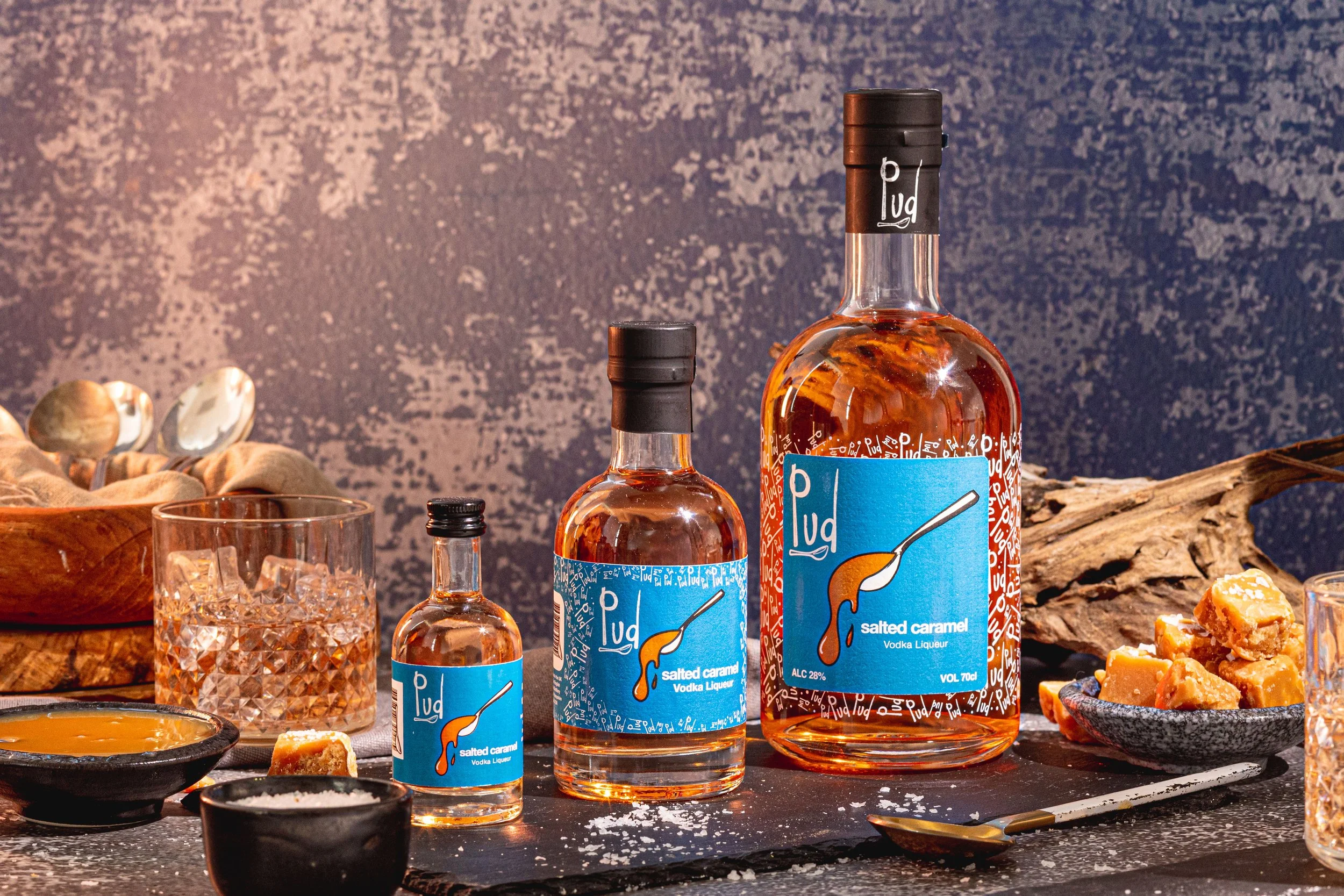 Display of three bottles of Pud salted caramel vodka liqueur, a glass of ice, small bowls with caramel sauce, whipped cream, and caramel fudge, on a dark surface with a textured dark background.