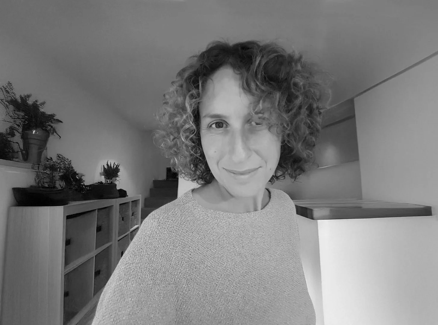 A woman with curly hair smiling and winking at the camera in a home interior with plants and furniture.