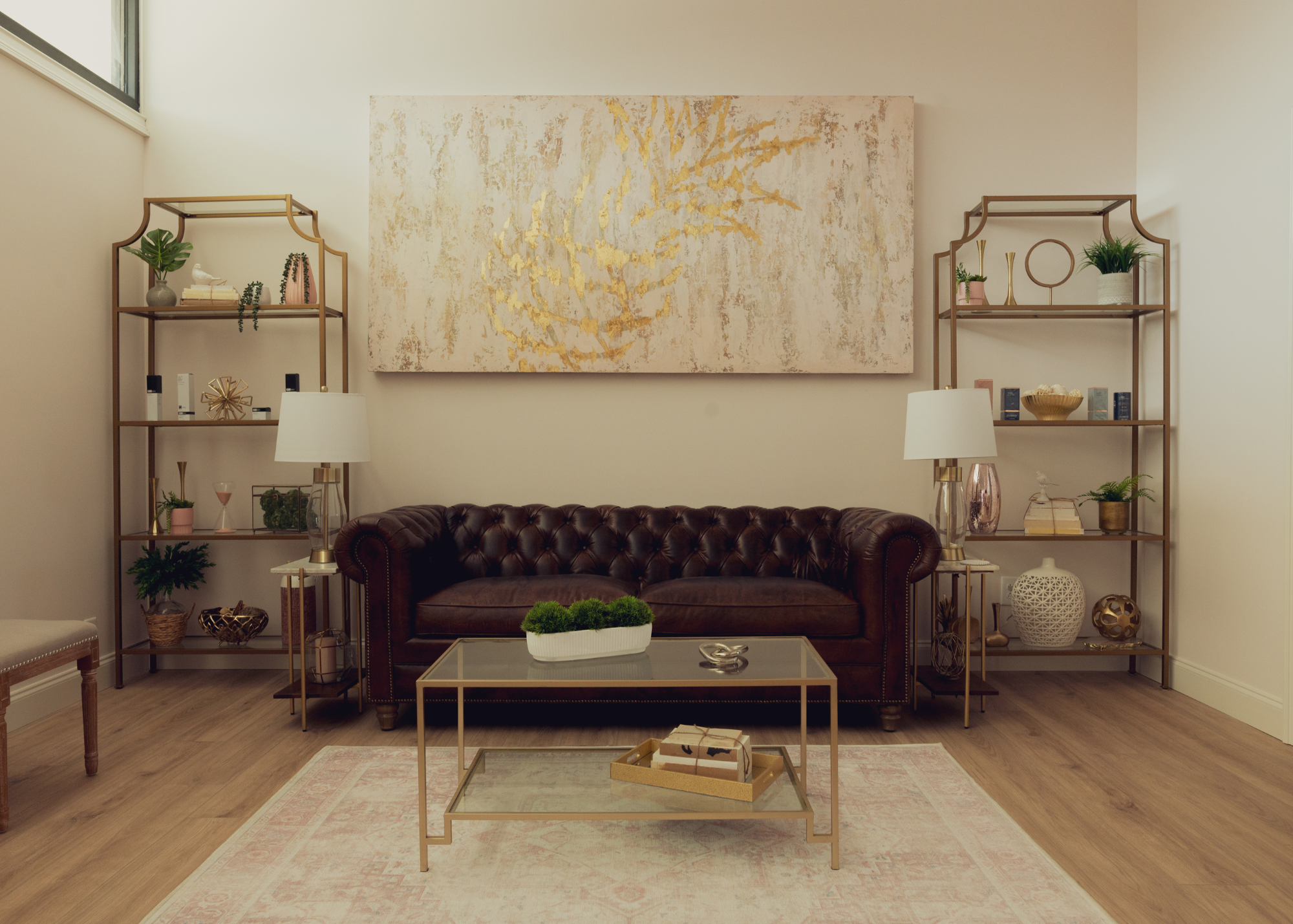 Living room with beige walls, a large abstract wall art with gold accents, a brown leather tufted sofa, two gold side tables with lamps, and wall-mounted shelves with decorative objects and plants.