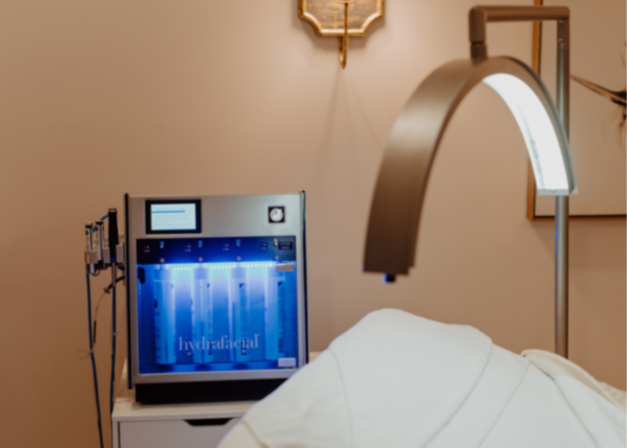 Medical device for hydrafacial treatment in a clinical setting, with a screen displaying instructions, next to a white bed with a patient lying down, and a nearby wall-mounted lamp.