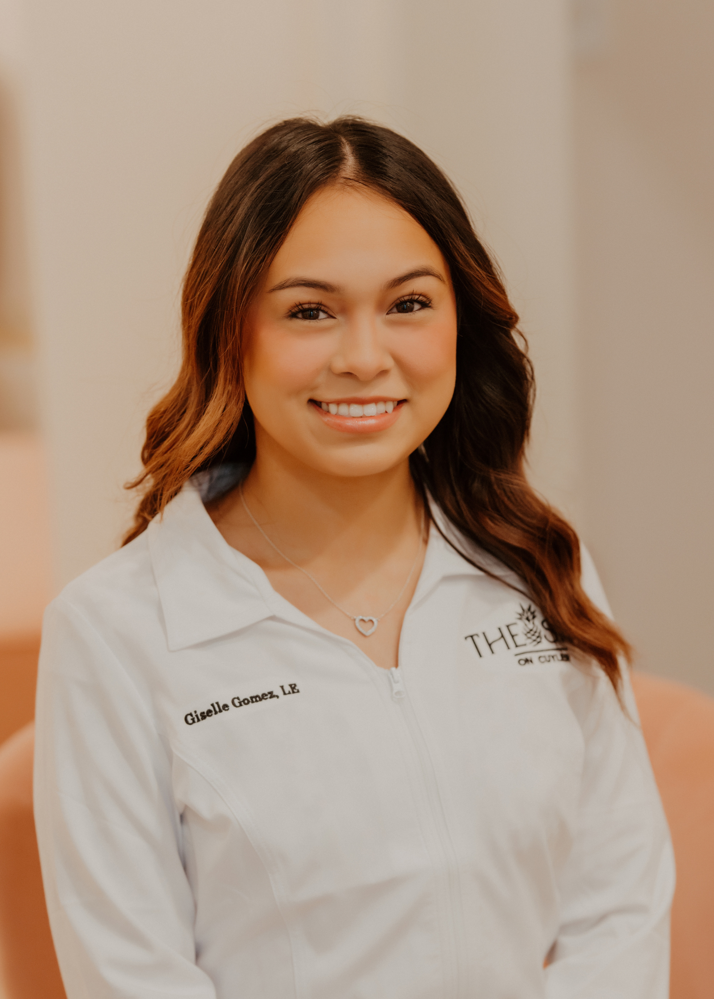 A young woman with dark wavy hair smiling, wearing a white zip-up jacket with her name and title embroidered on it, and a heart-shaped necklace.