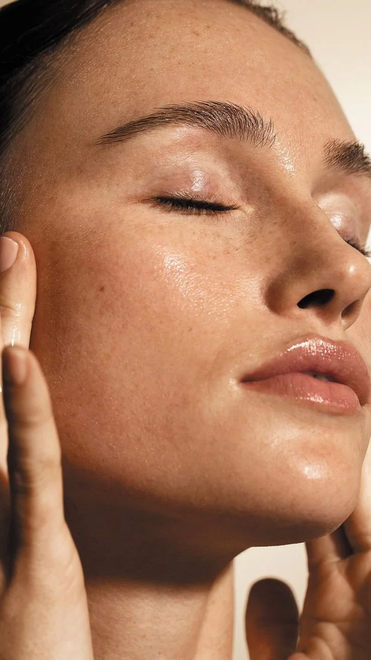 Close-up of a woman's face with clear, glowing skin, eyes closed, and hands gently touching her cheeks.
