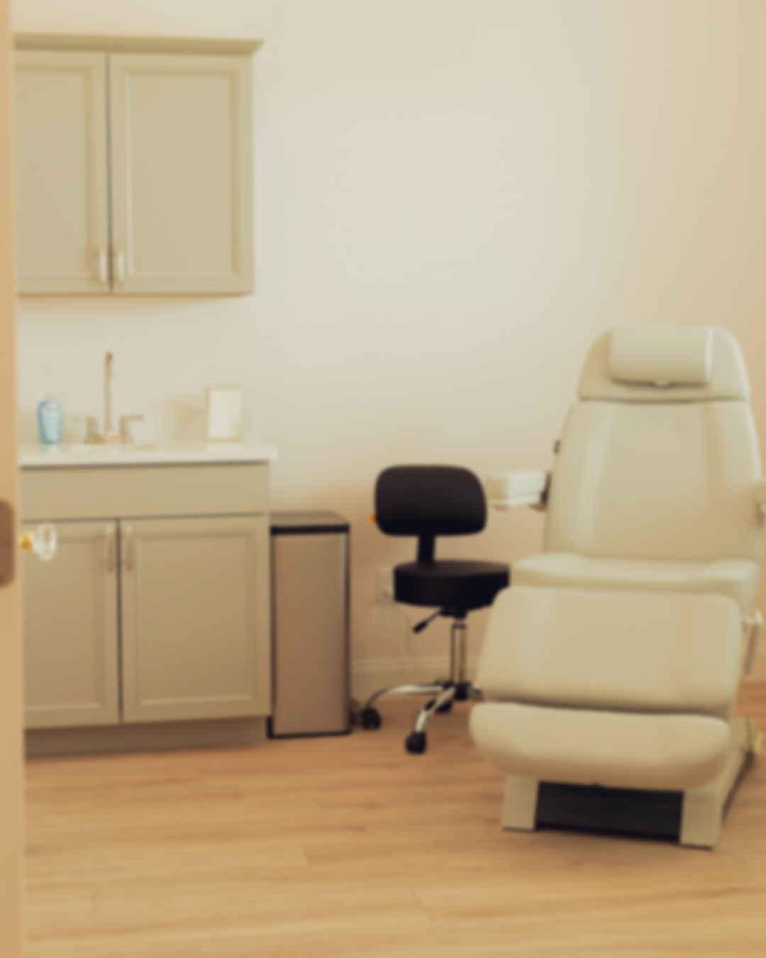 Medical examination room with a white treatment chair, a black stool, a counter with hand sanitizer, a small refrigerator, and a cabinet.