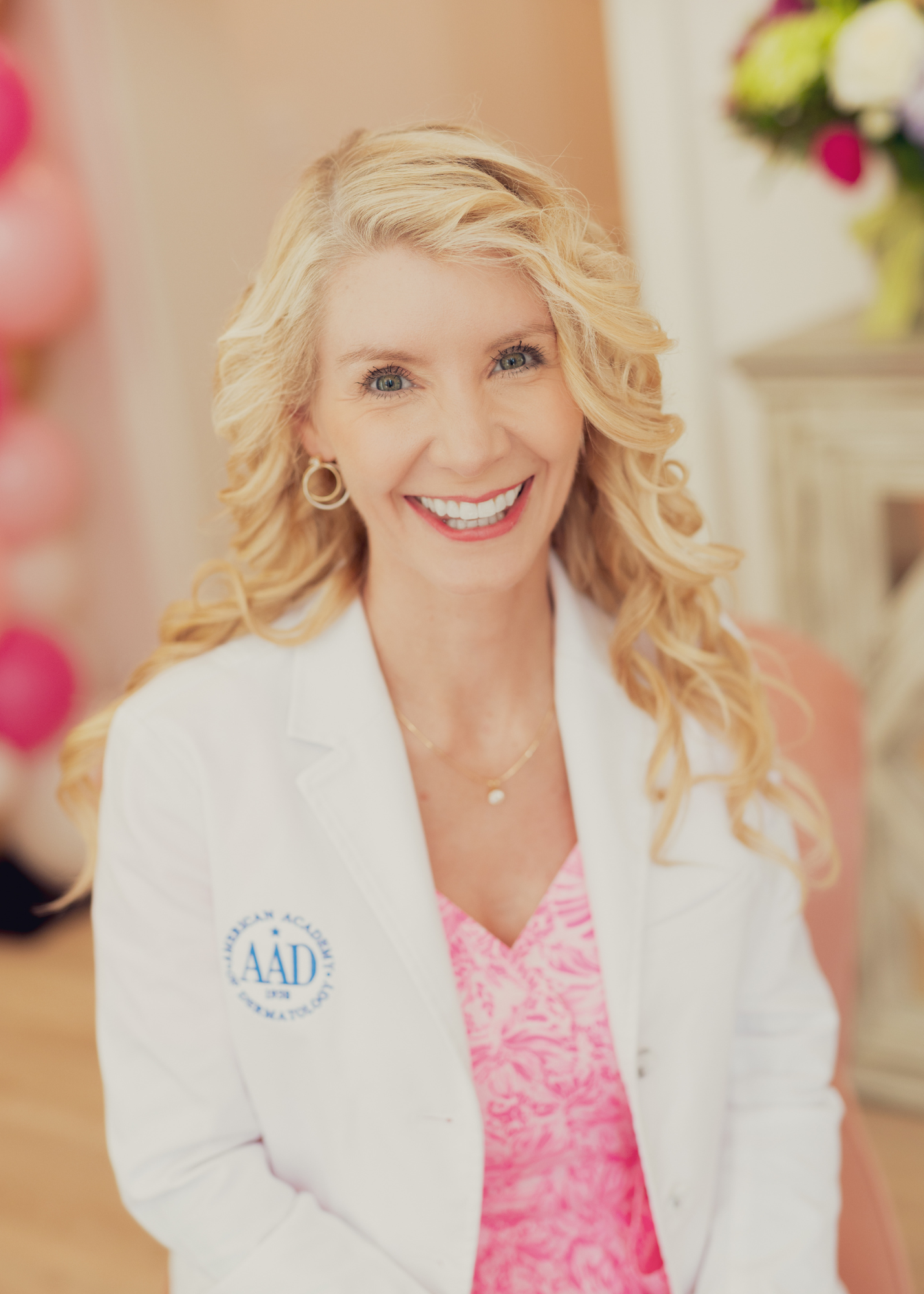 Woman with blonde curly hair and blue eyes smiling, wearing a white blazer with a logo, pink patterned shirt, and gold jewelry, standing indoors with blurred floral background.