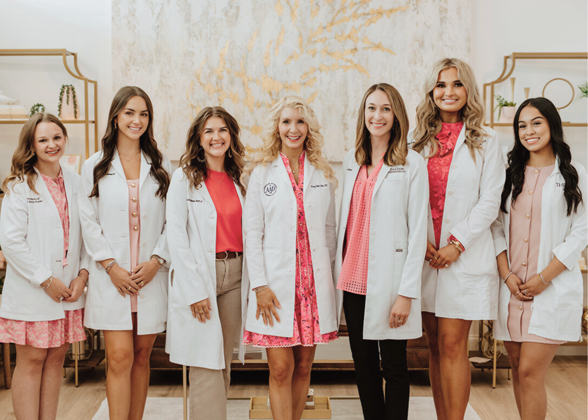 Group of seven women in white coats standing together in a bright room with light-colored walls and shelves with plants and decor.