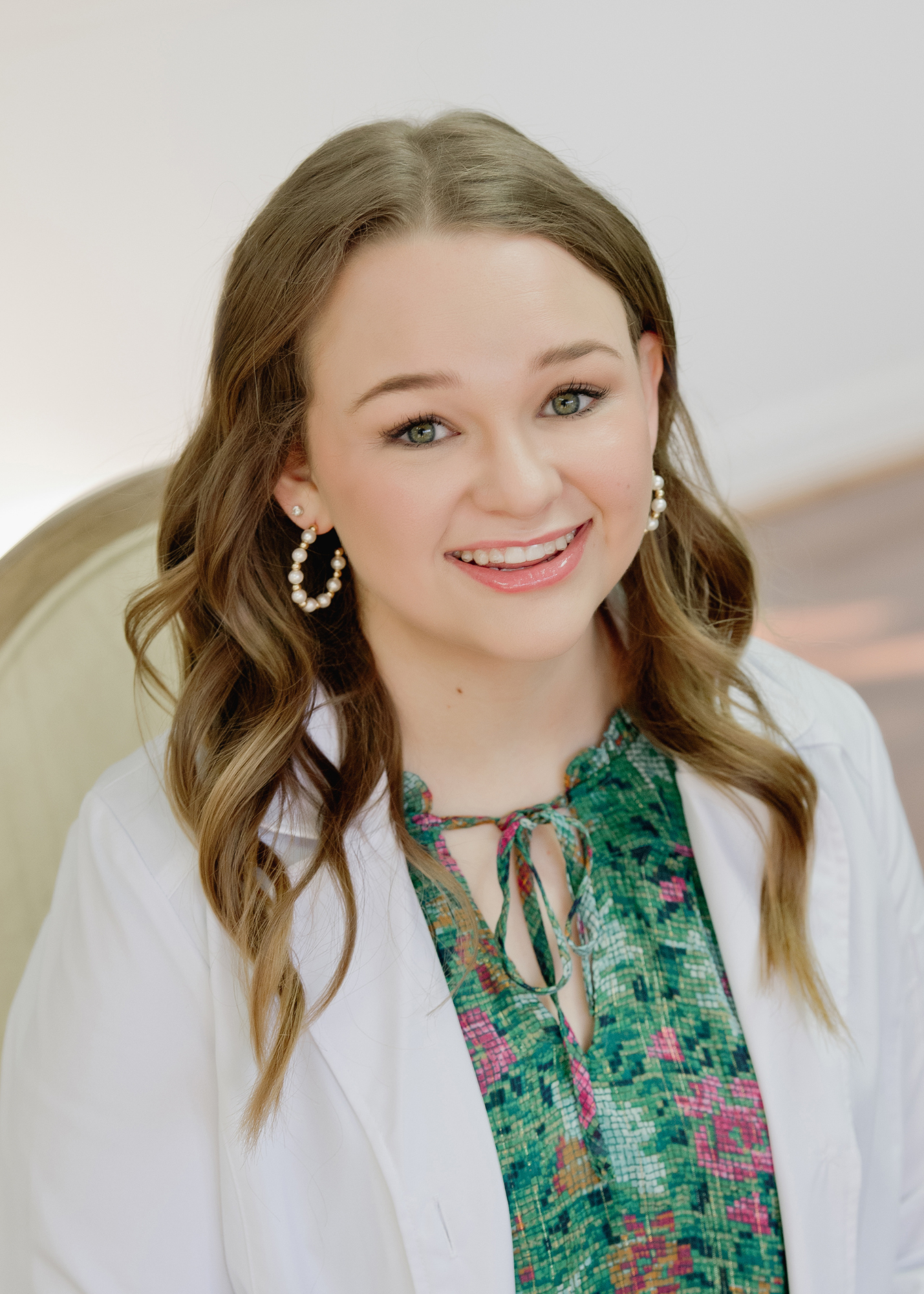 Young woman with long wavy brown hair smiling, wearing pearl earrings and a colorful green patterned top with a white jacket, sitting in a cream-colored chair against a plain light background.