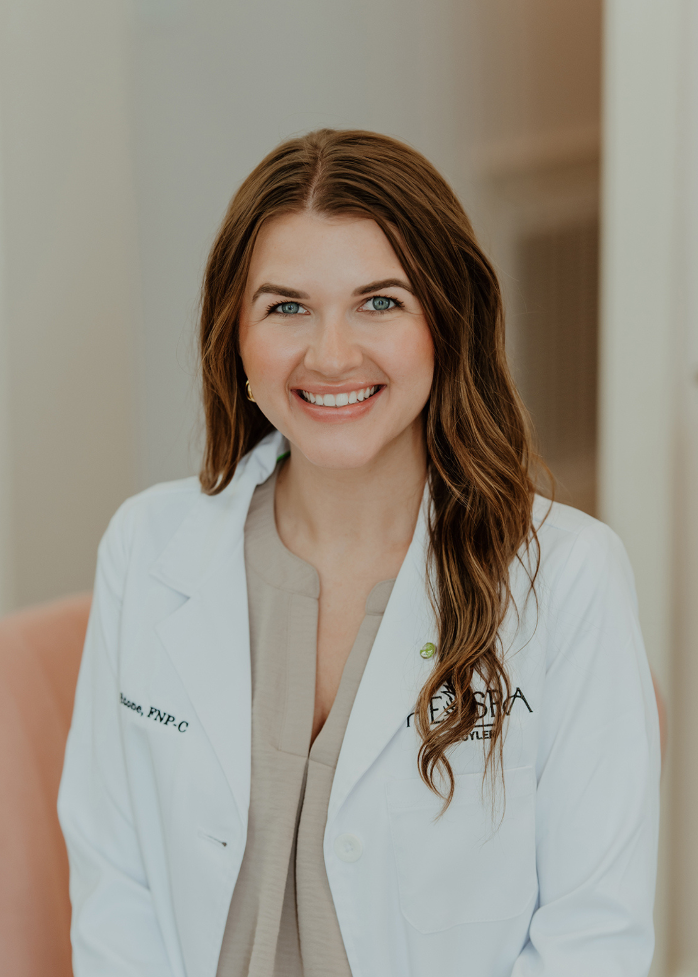 A smiling woman with long, wavy brown hair wearing a white lab coat and beige top in a medical or clinical setting.