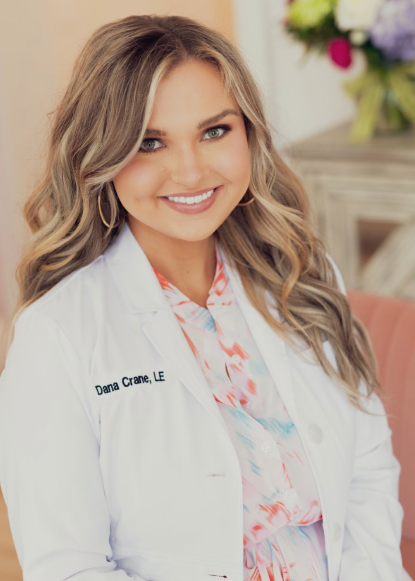 A smiling woman wearing a white lab coat with a name tag that reads 'Dana Crane, LE', and a colorful blouse with a floral pattern. Behind her are blurred flowers.