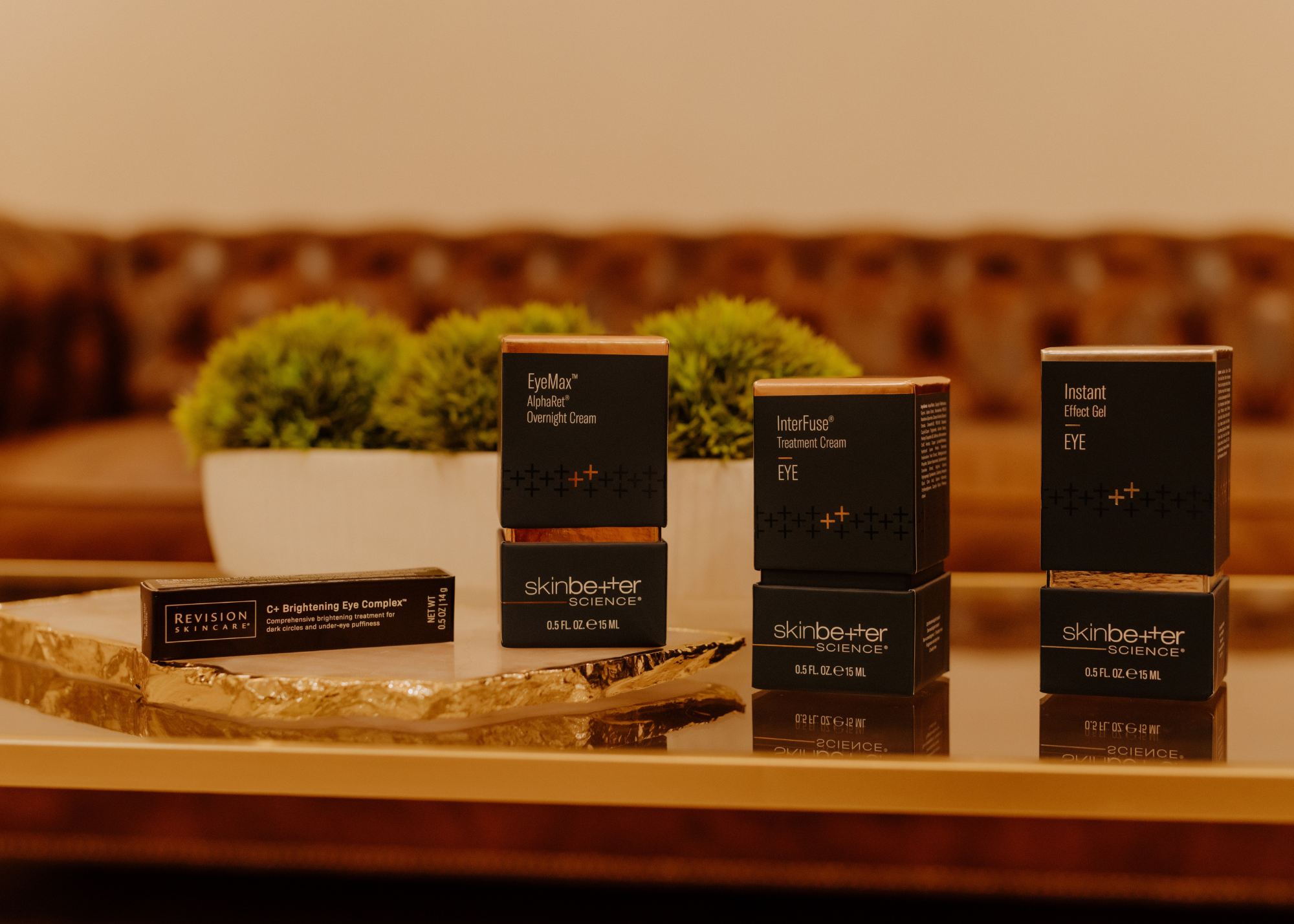 A collection of skincare products including EyeMax Overnight Cream, InterFuse Treatment Cream, and Instant Effect Gel for eyes, displayed on a wooden surface with a blurred background of green plants and a beige wall.