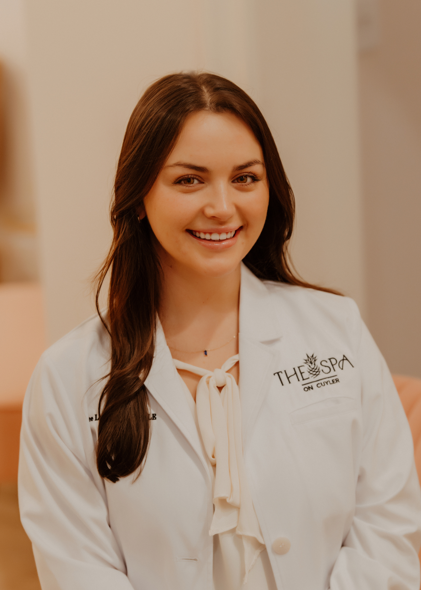 A smiling woman with long brown hair wearing a white coat with a logo, standing indoors.