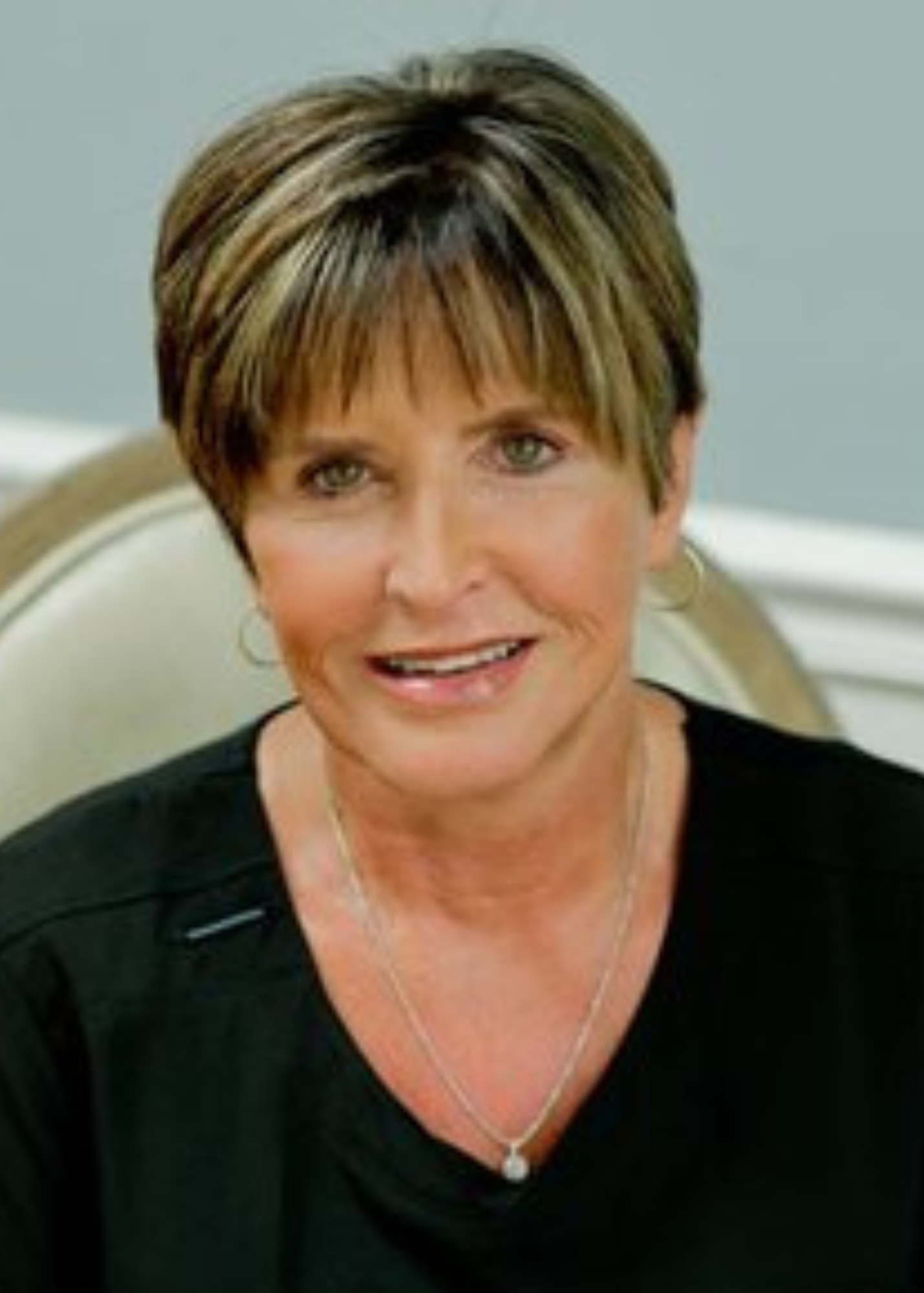 Close-up of a woman with short, layered, brown hair with blonde highlights, wearing a black top and silver jewelry, smiling in an indoor setting.