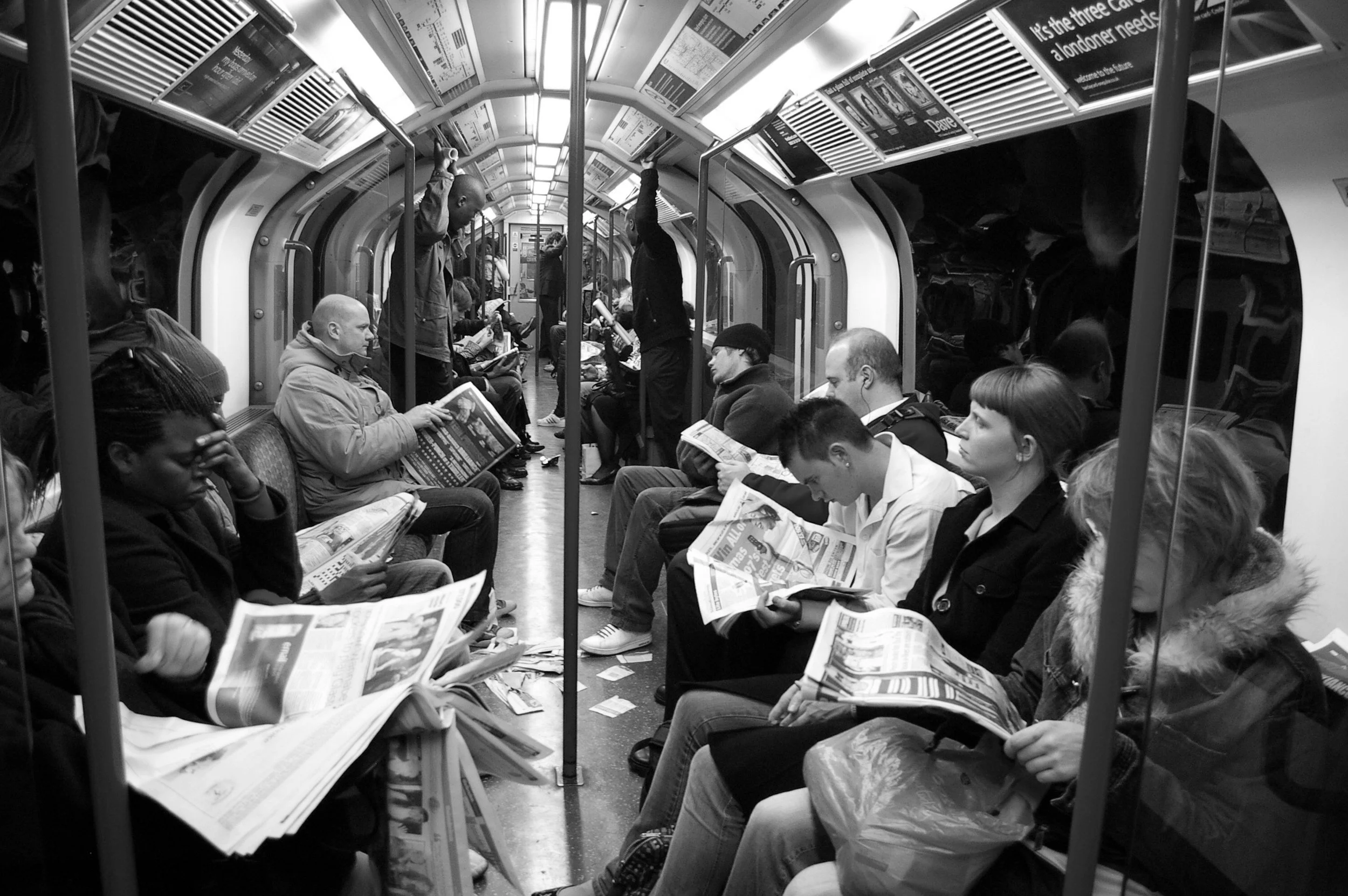 People sitting and standing inside a subway car, many reading newspapers or looking at their phones, with some holding onto the overhead handrails.