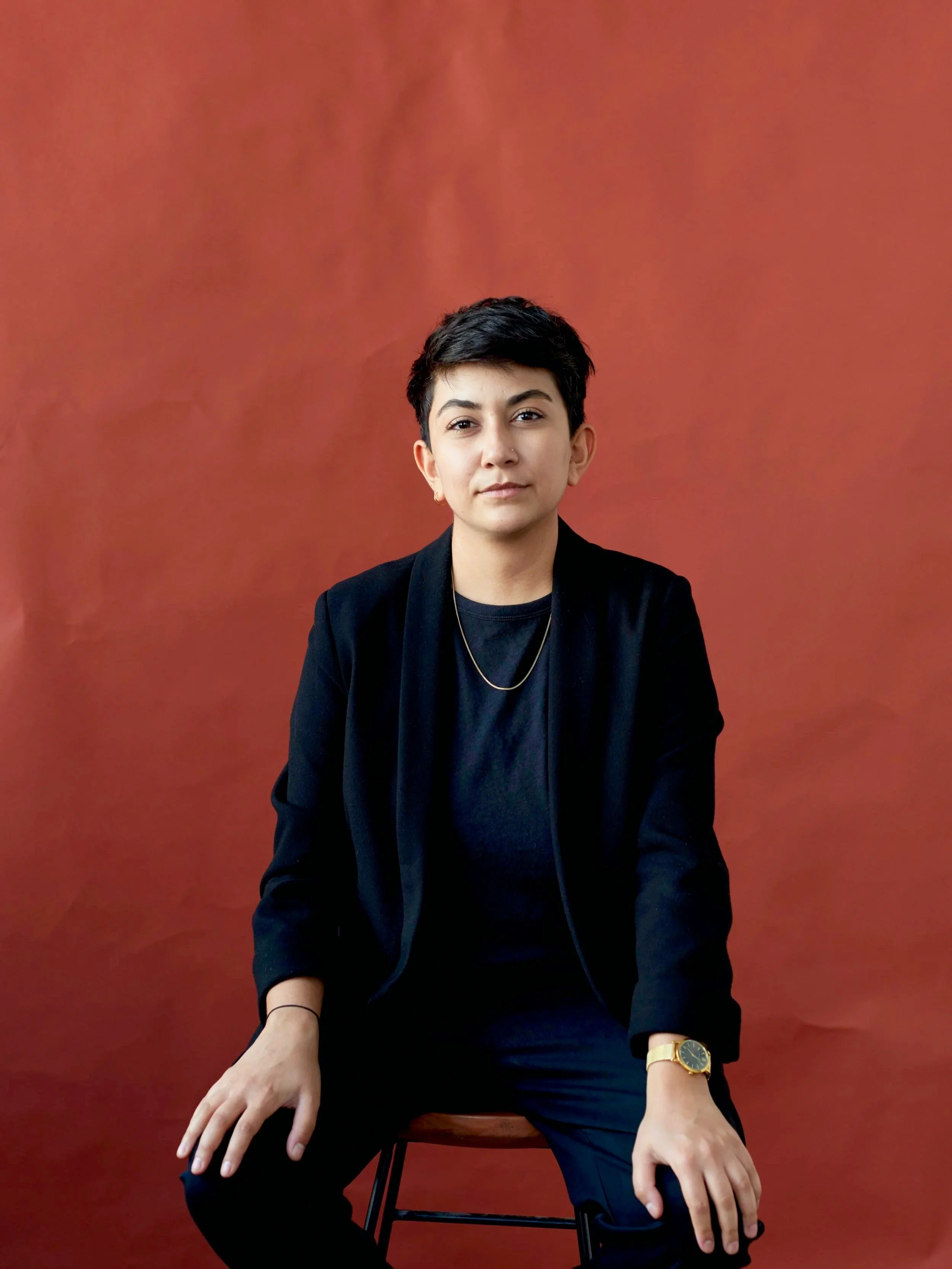 Emissary Founder, Meera Pattni, wearing a black blazer, black shirt, and gold watch, seated on a stool against a textured red background.