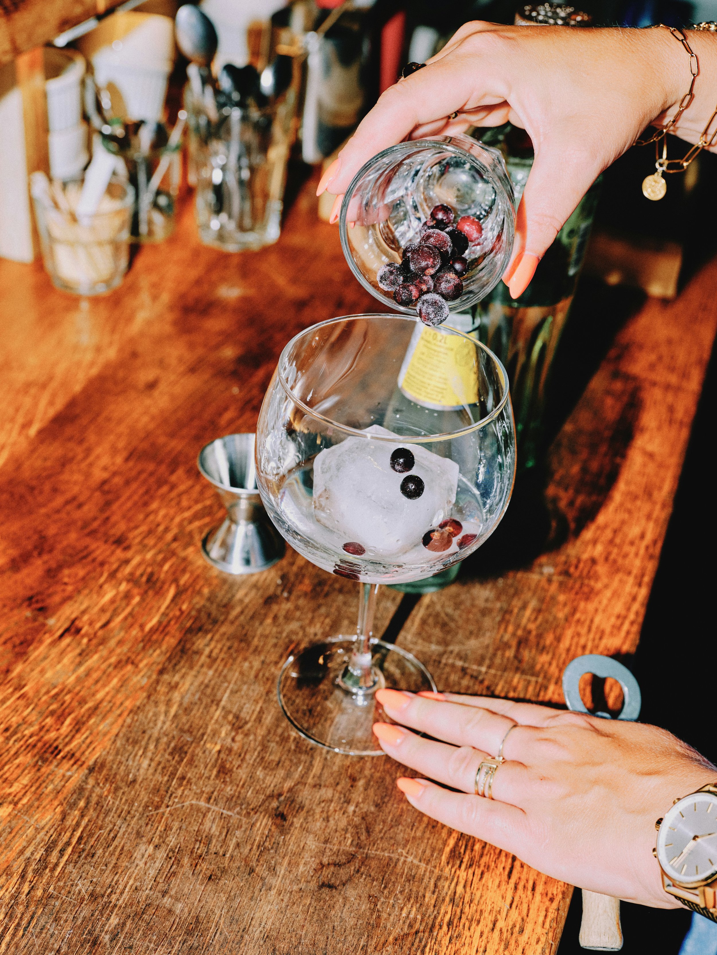 A person pouring frozen berries into a large cocktail glass with ice, on a wooden bar counter.