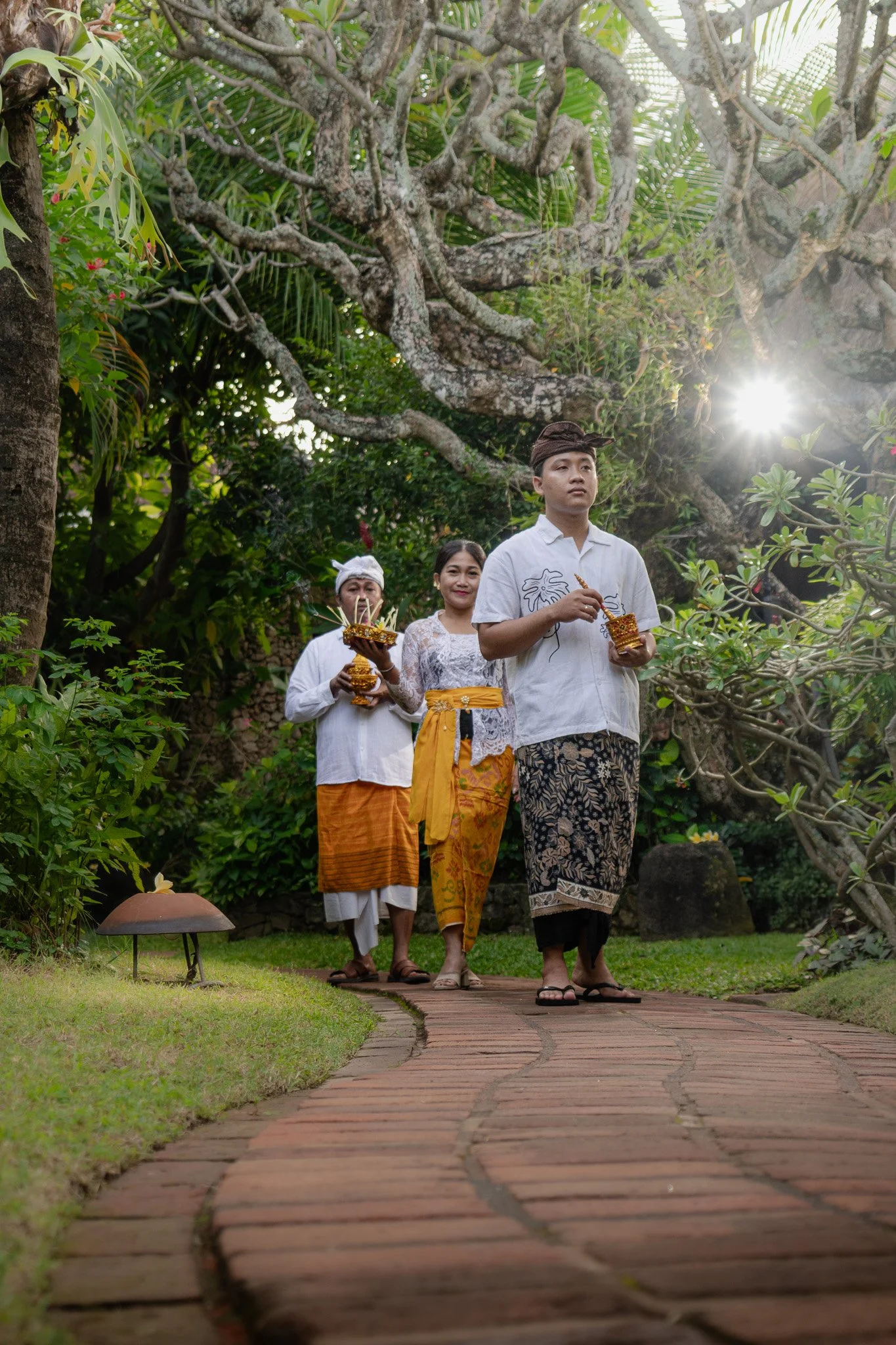 Two men and a woman walk down a path in Poppies Hotel gardens holding spiritual offerings as part of a procession for Balinese Hinduism