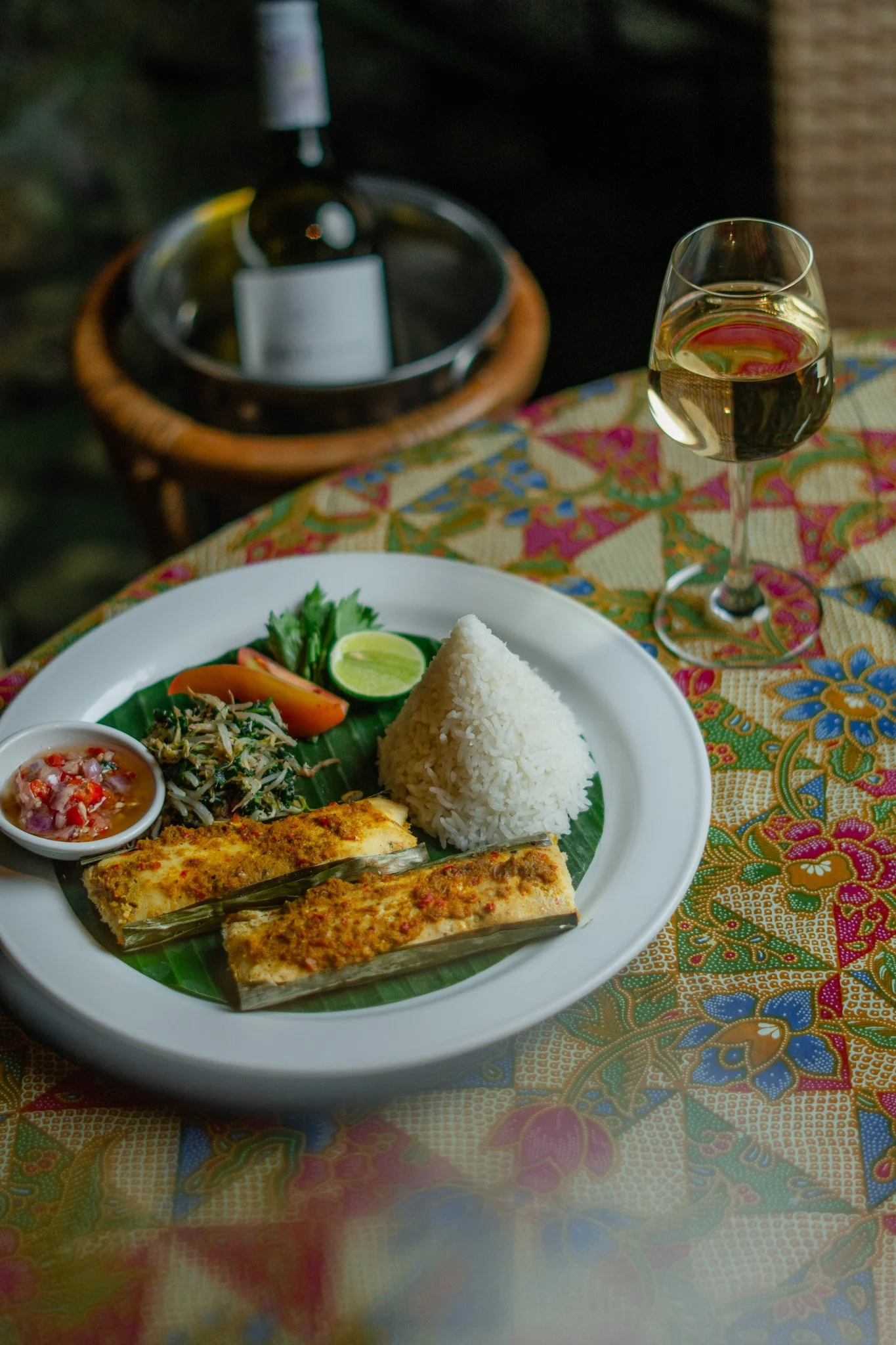 Traditional Indonesian dish called Ikan Pepes, featuring fish seasoned with spices, wrapped in banana leaves, and grilled. A glass of wine sits on the table cloth.