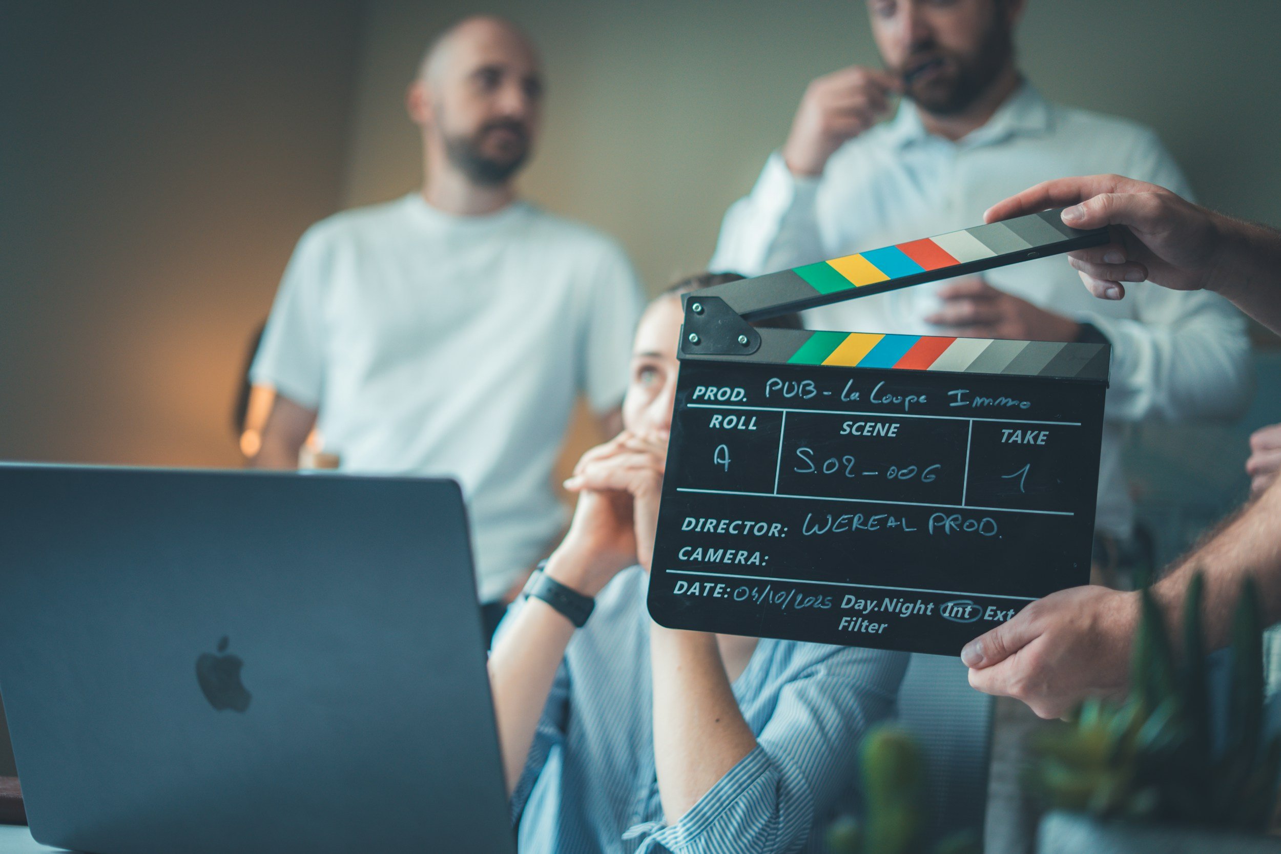 Behind the scenes of a film production, a film slate is held in front of a woman sitting at a laptop while two men stand behind her in discussion.