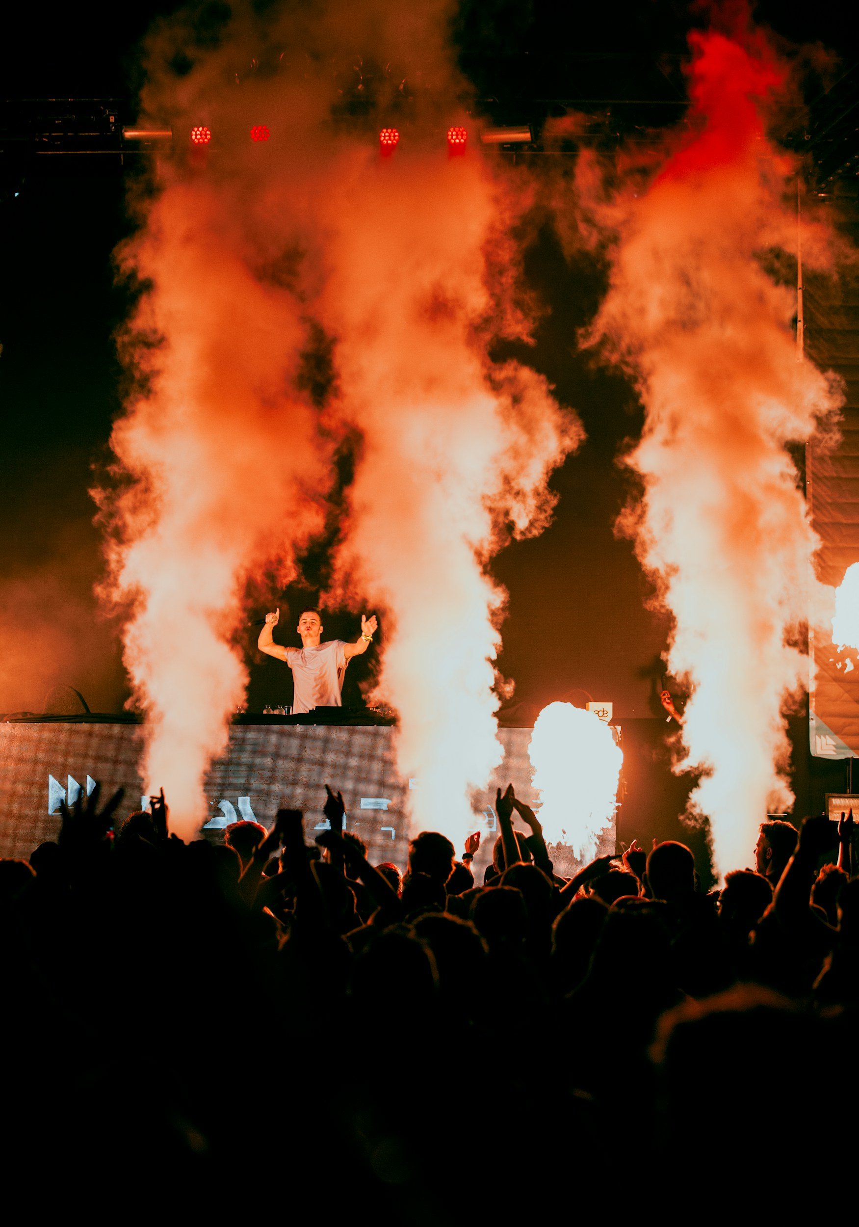 A DJ performs on stage with large fire and smoke effects, crowd dancing in front at a concert or music festival.