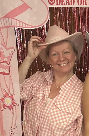 A woman smiling and wearing a cowboy hat, pink checkered shirt, standing in front of a pink sequin backdrop with game signs and decorations.