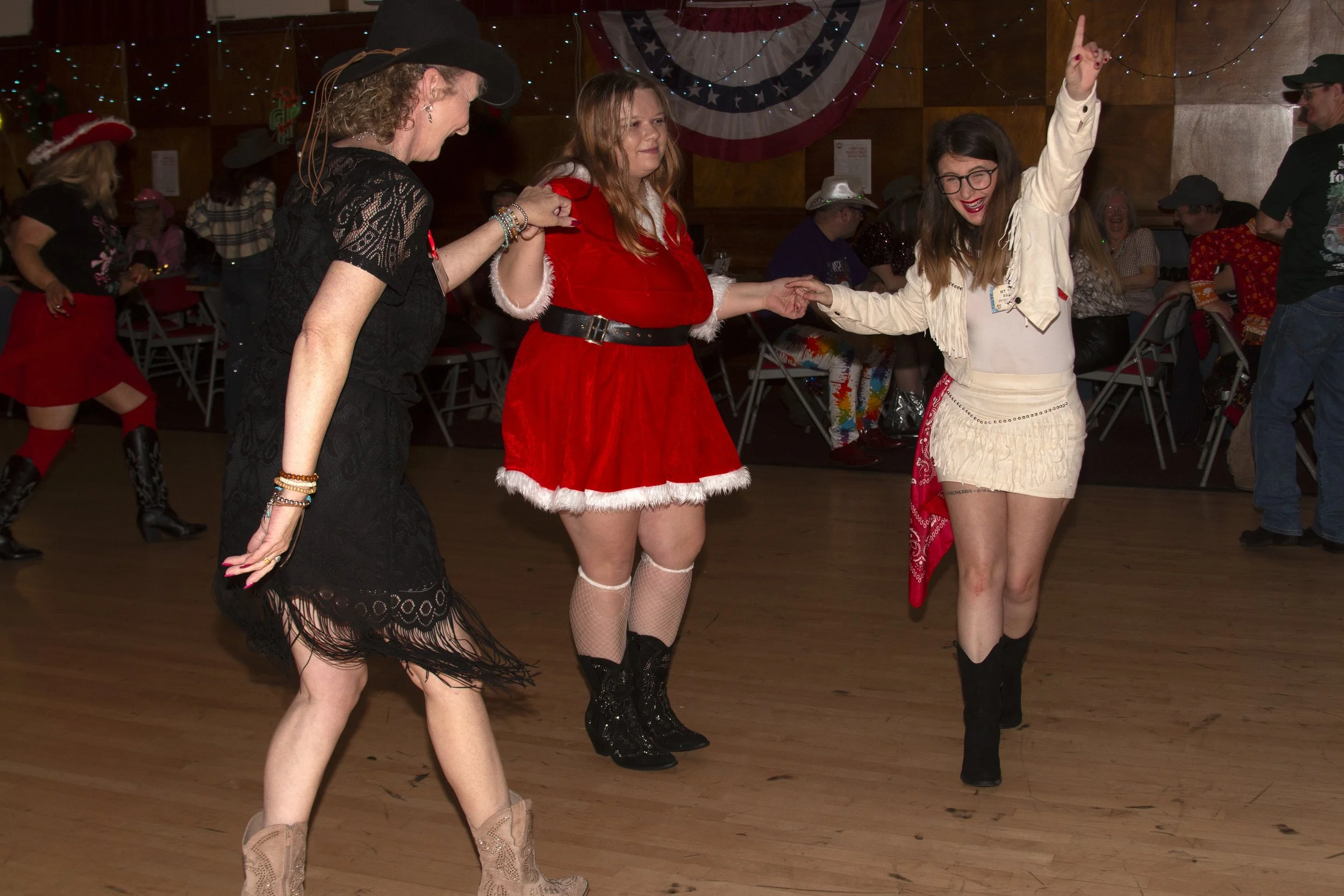 Three women dancing at a holiday-themed party, with some dressed in festive costumes. The woman in the center wears a red Santa dress, while the woman on the right wears a white western outfit with black boots. The woman on the left wears a black lace dress and cowboy boots. The background has Christmas decorations and other guests.